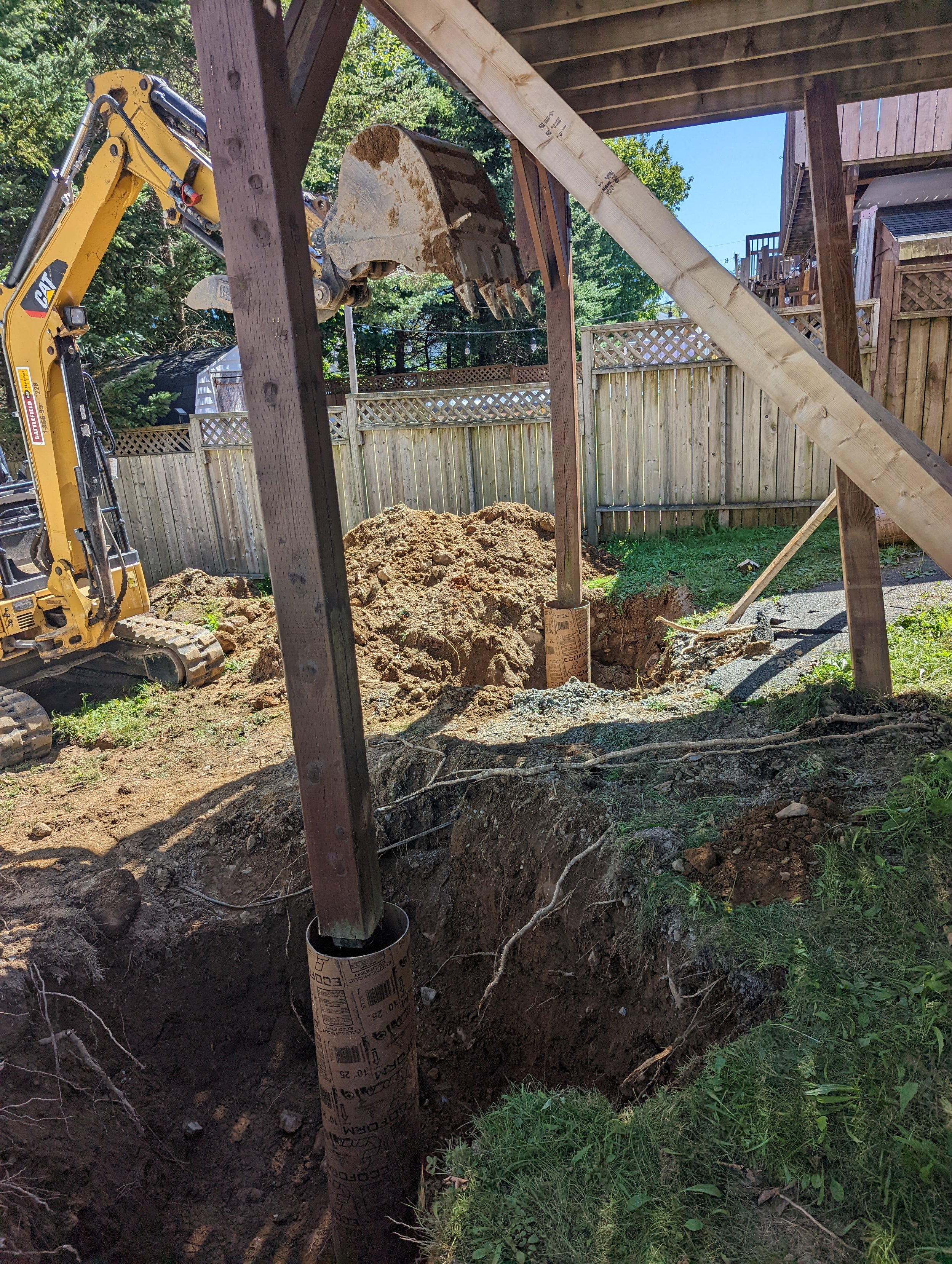 Backyard construction site with excavator digging holes and supporting posts, surrounded by dirt and wooden fence.