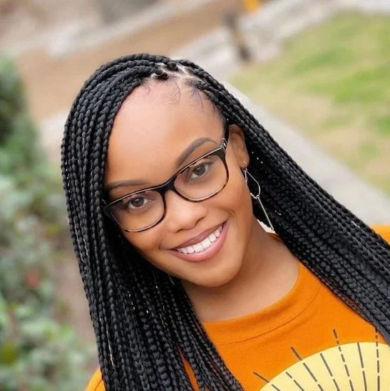 A young woman with long black braided hair, glasses, and hoop earrings smiling outdoors in autumn weather.