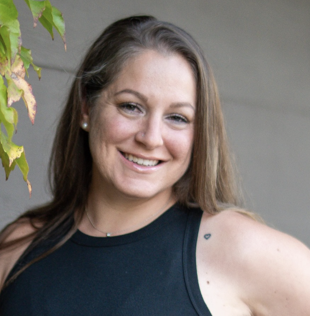 Smiling young woman with long brown hair, wearing a black sleeveless top, standing outdoors near a gray wall and some green leaves.