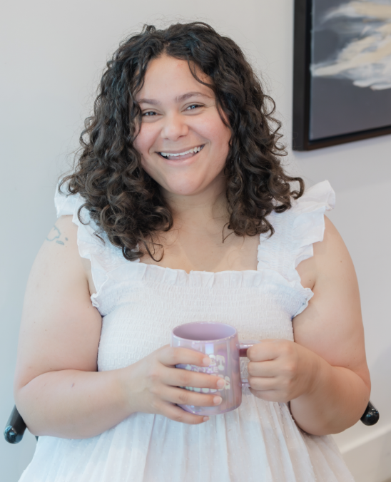 A woman with curly dark hair smiling while holding a purple mug.