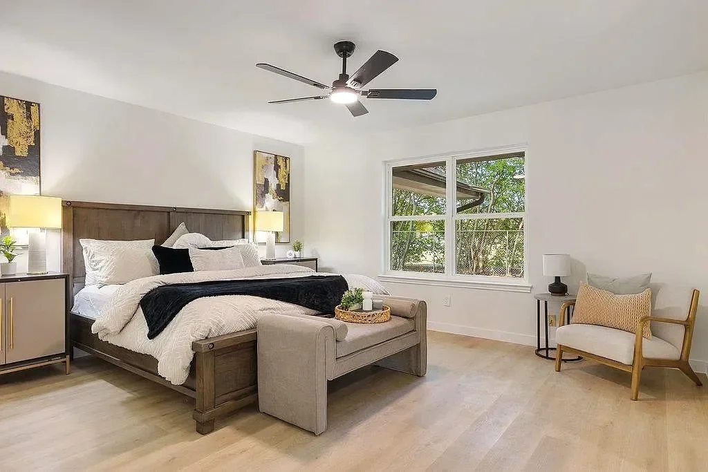 A spacious bedroom with white walls, a wooden bed frame, white bedding, black and white accent pillows, two matching nightstands with lamps, a ceiling fan, a beige chaise lounge at the foot of the bed, a window showing greenery outside, and a white armchair with a light-colored cushion beside a small side table.