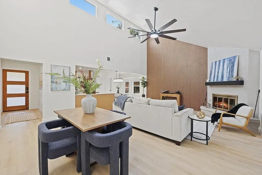 Modern living room with white sofa, armchair, fireplace, and wooden accent wall, high ceiling with ceiling fan, light wooden flooring, and large windows.