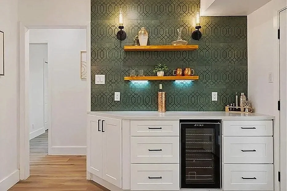 A bar area with white cabinetry, a built-in wine cooler, and floating wooden shelves mounted on a dark patterned accent wall. The shelves hold decorative items including a vase, potted plant, and mugs. There are sconces providing ambient lighting.