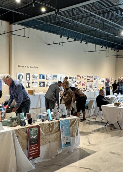 People browsing photo exhibition with tables displaying books and photographs on the wall.