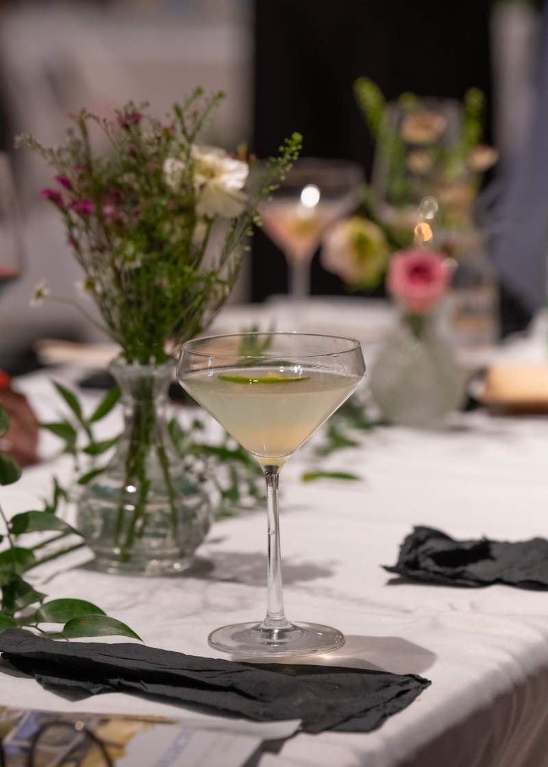 A close-up of a cocktail glass with a light-colored drink and lime garnish on a white tablecloth, with additional flowers and drinks in the blurred background.