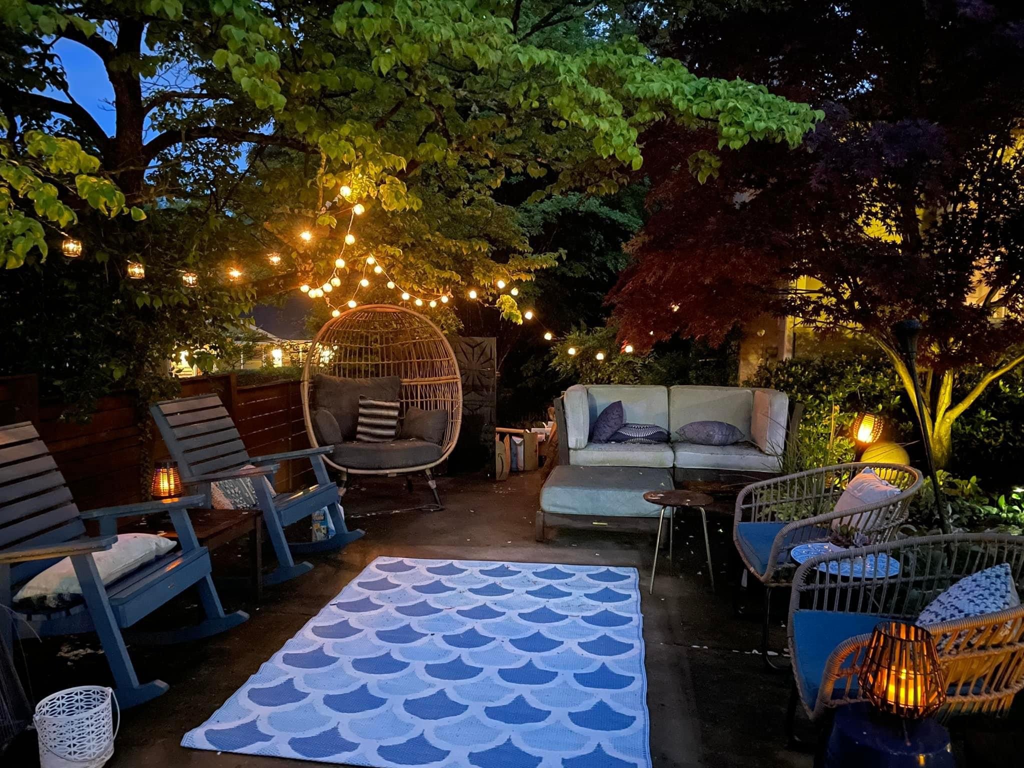An outdoor patio at dusk with string lights, a hanging egg chair with cushions, sofas with pillows, wicker chairs, small tables with lanterns, and a patterned rug underfoot, surrounded by lush trees.
