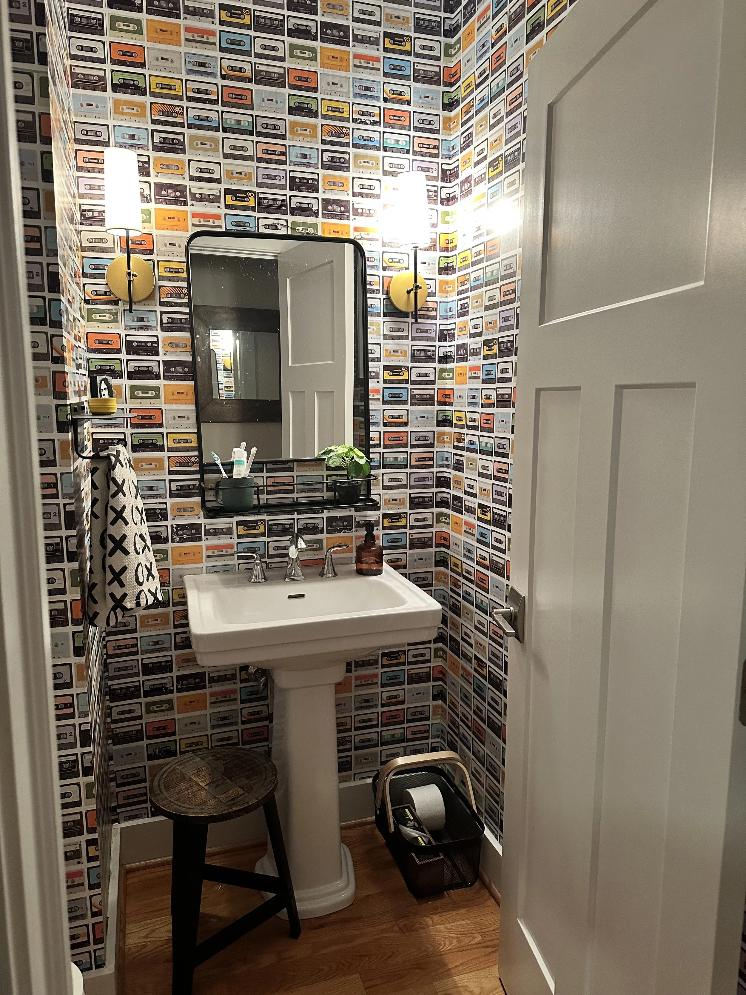 Small bathroom with vintage cassette tape wallpaper, a white sink with a mirror above, two wall sconces, a towel hanging on a rack, a small wooden stool, and a trash bin with a paper towel roll.
