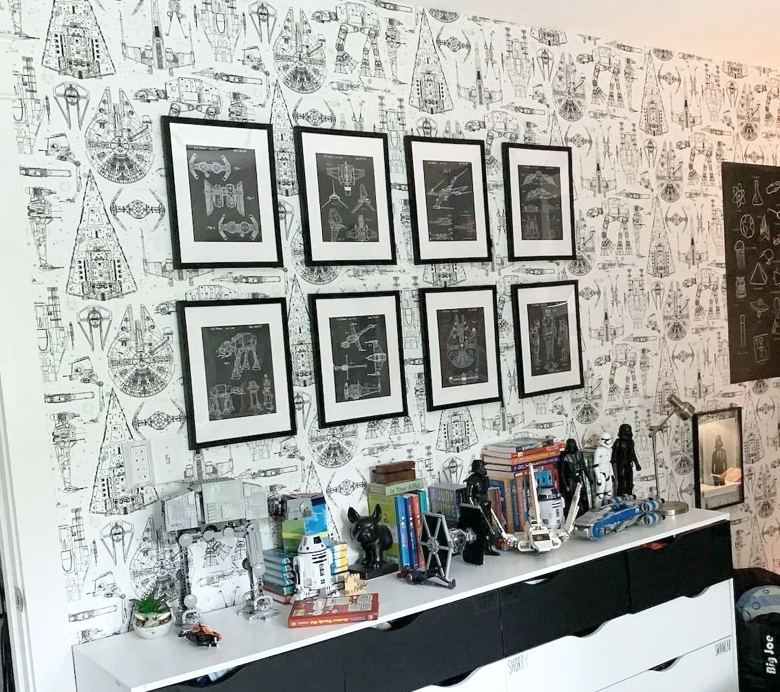 A wall decorated with black and white space and starship sketches, with five framed space ship blueprints hanging above a white dresser. The dresser holds Star Wars toys, books, and figurines, including R2-D2 and Darth Vader.