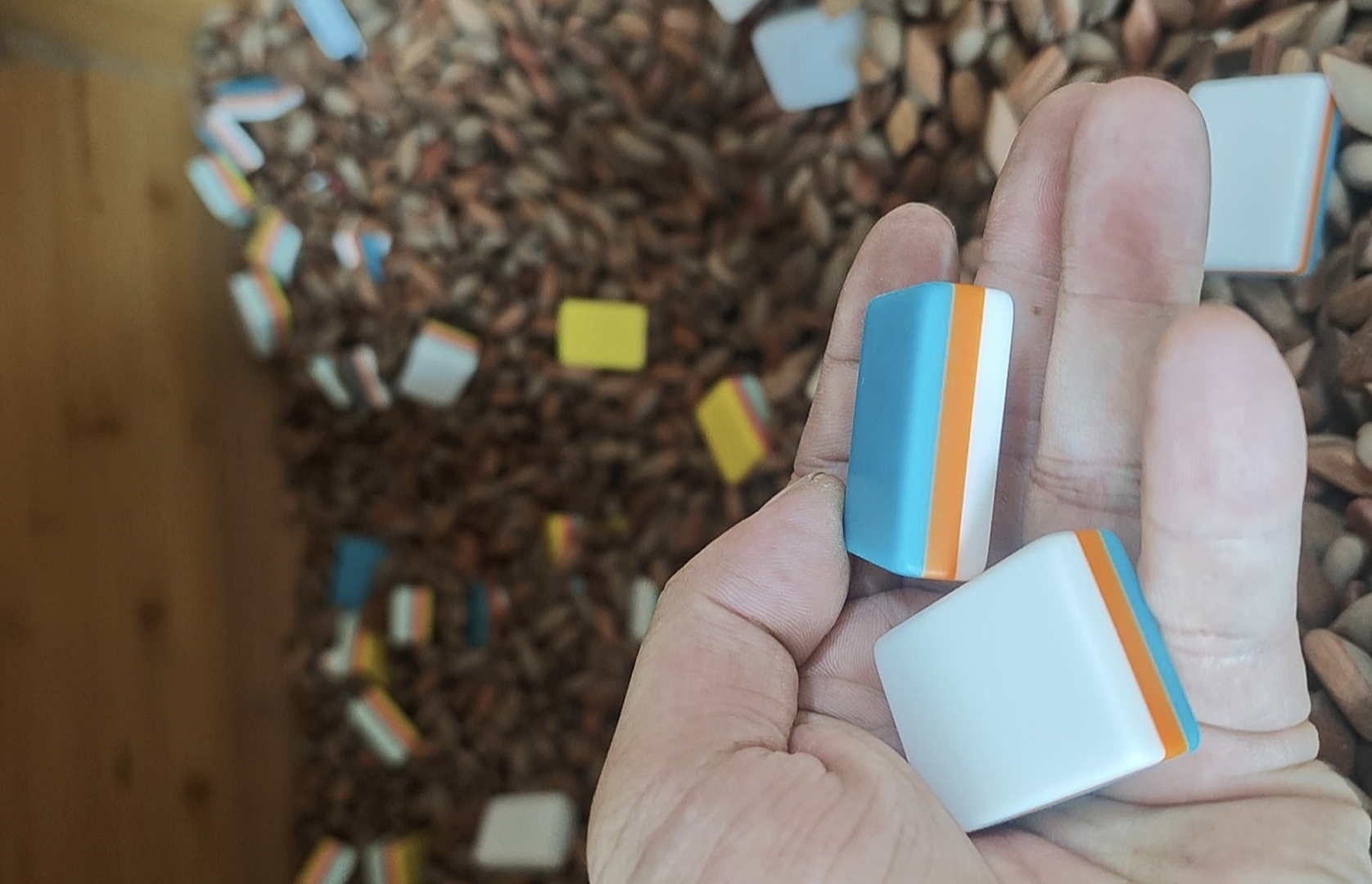A hand holding two mahjong tiles with no design, with layers of teal, orange, and white. The background shows the tumbling polishing material.