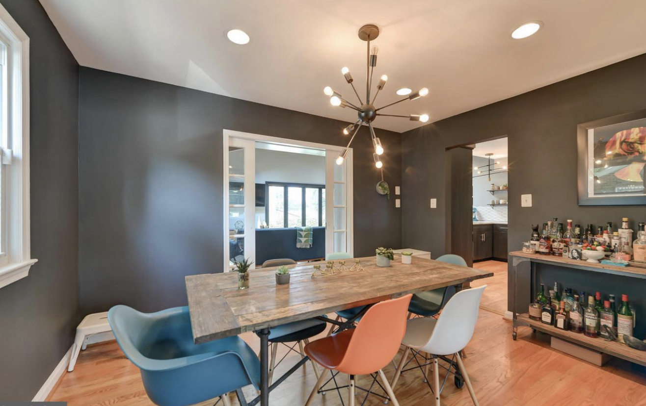 A dining room with a wooden table and five colorful chairs, gray walls, a modern chandelier, and a bar cart with alcohol bottles.