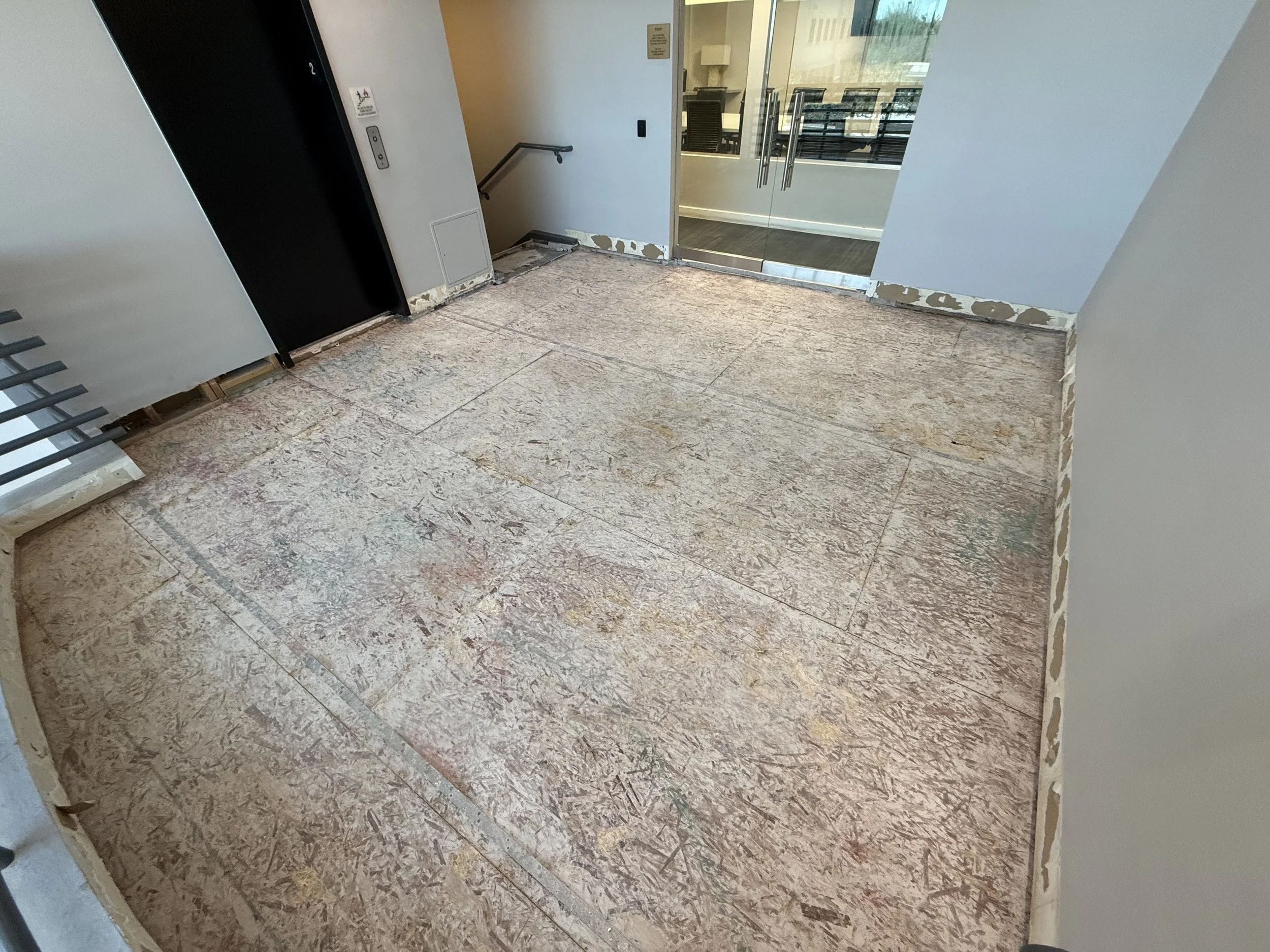 Indoor space under construction or renovation with unfinished flooring and partially finished walls, near an elevator and glass door leading to an outdoor area.