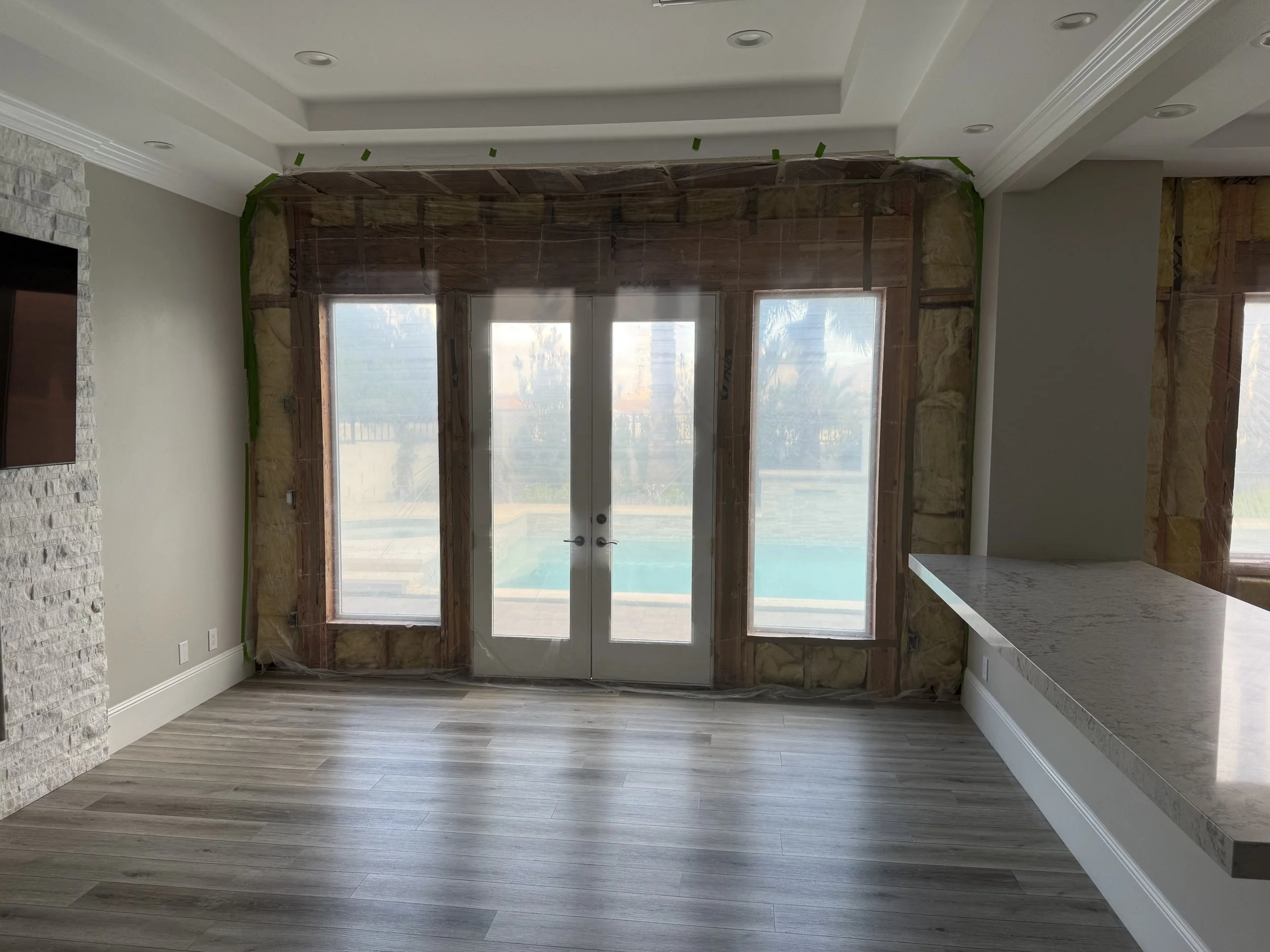 Interior of a room under construction with a view of a pool and yard outside through large windows and glass doors, with some wall insulation exposed.