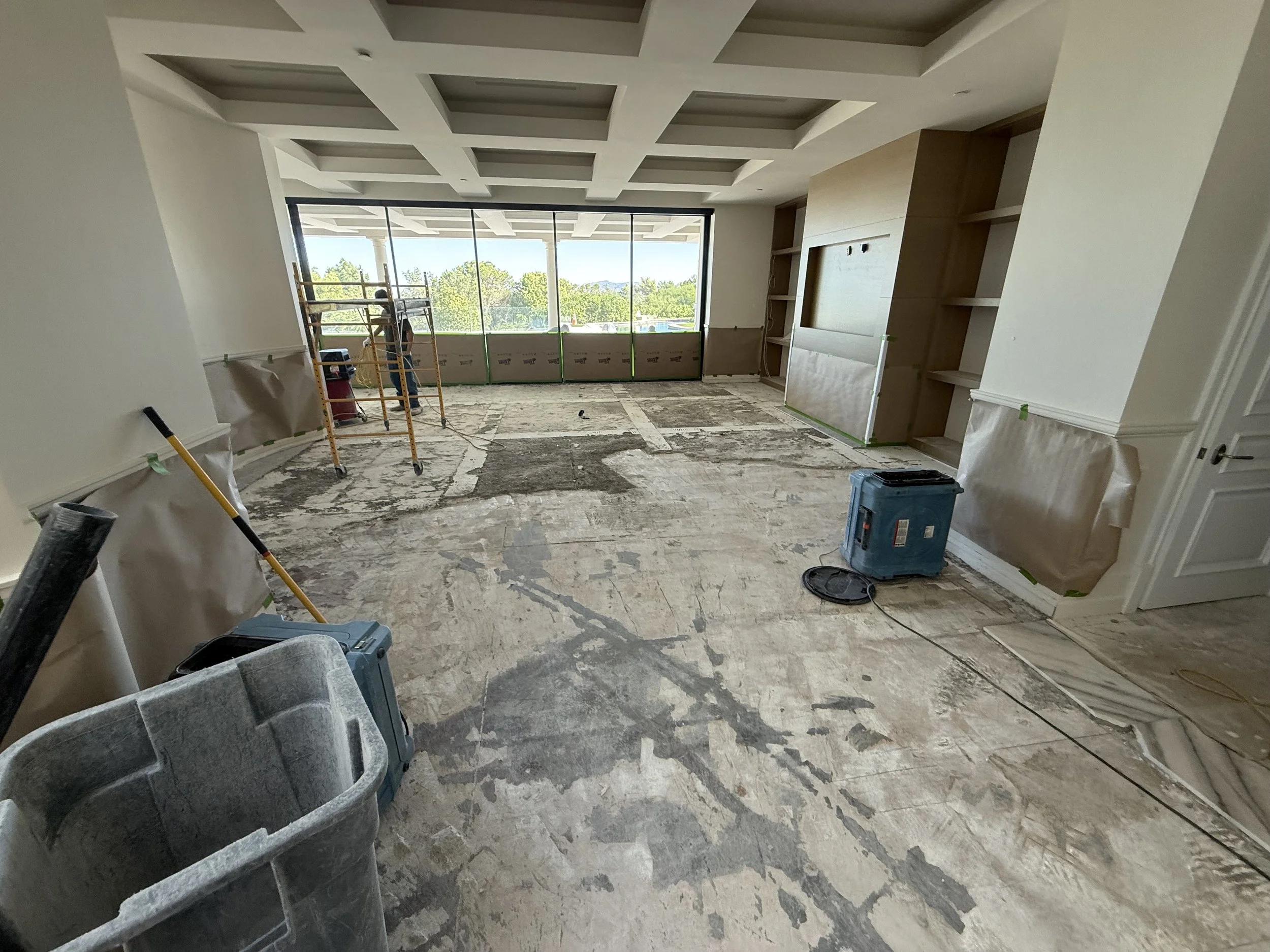 Living room under renovation with construction tools, scaffolding, and a large window view of trees outside.