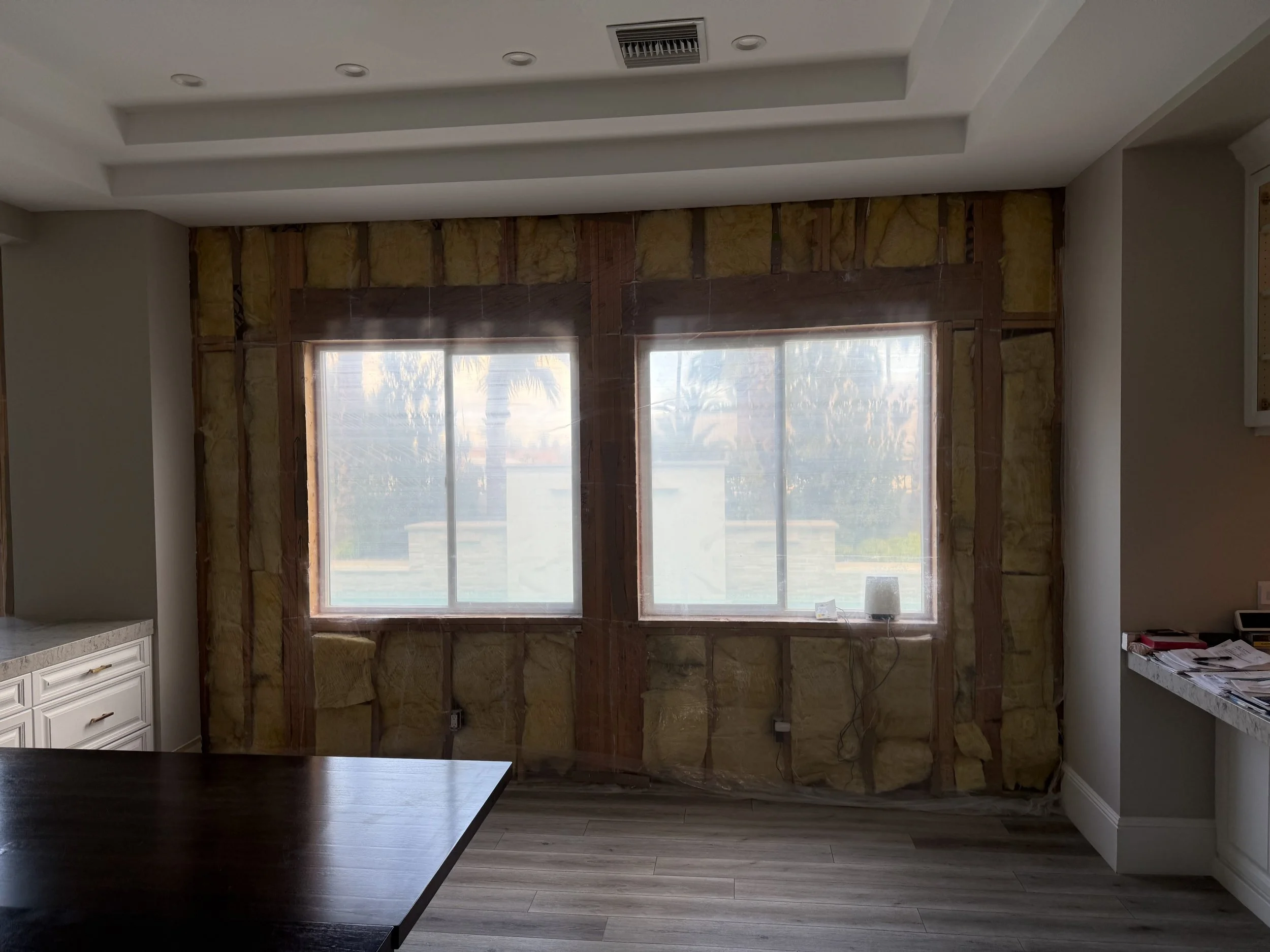 Kitchen undergoing renovation with exposed insulation and framing around two large windows, with a view of palm trees outside.