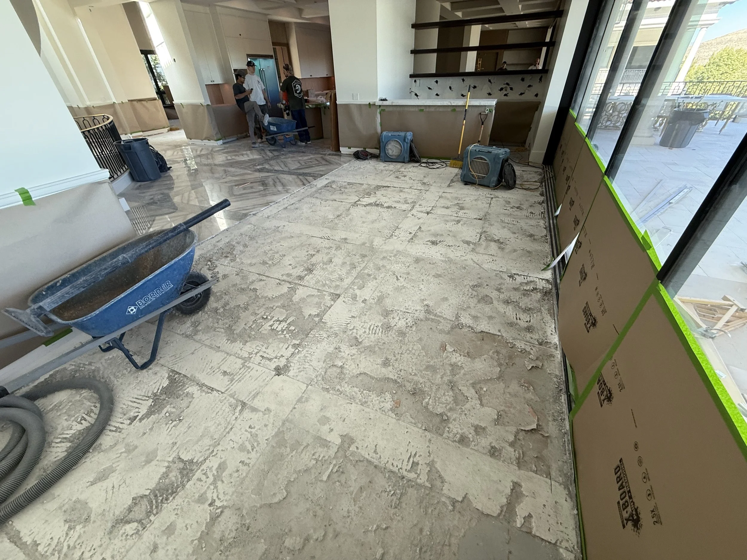Interior of a building under renovation with unfinished flooring, construction equipment, and workers in the background.
