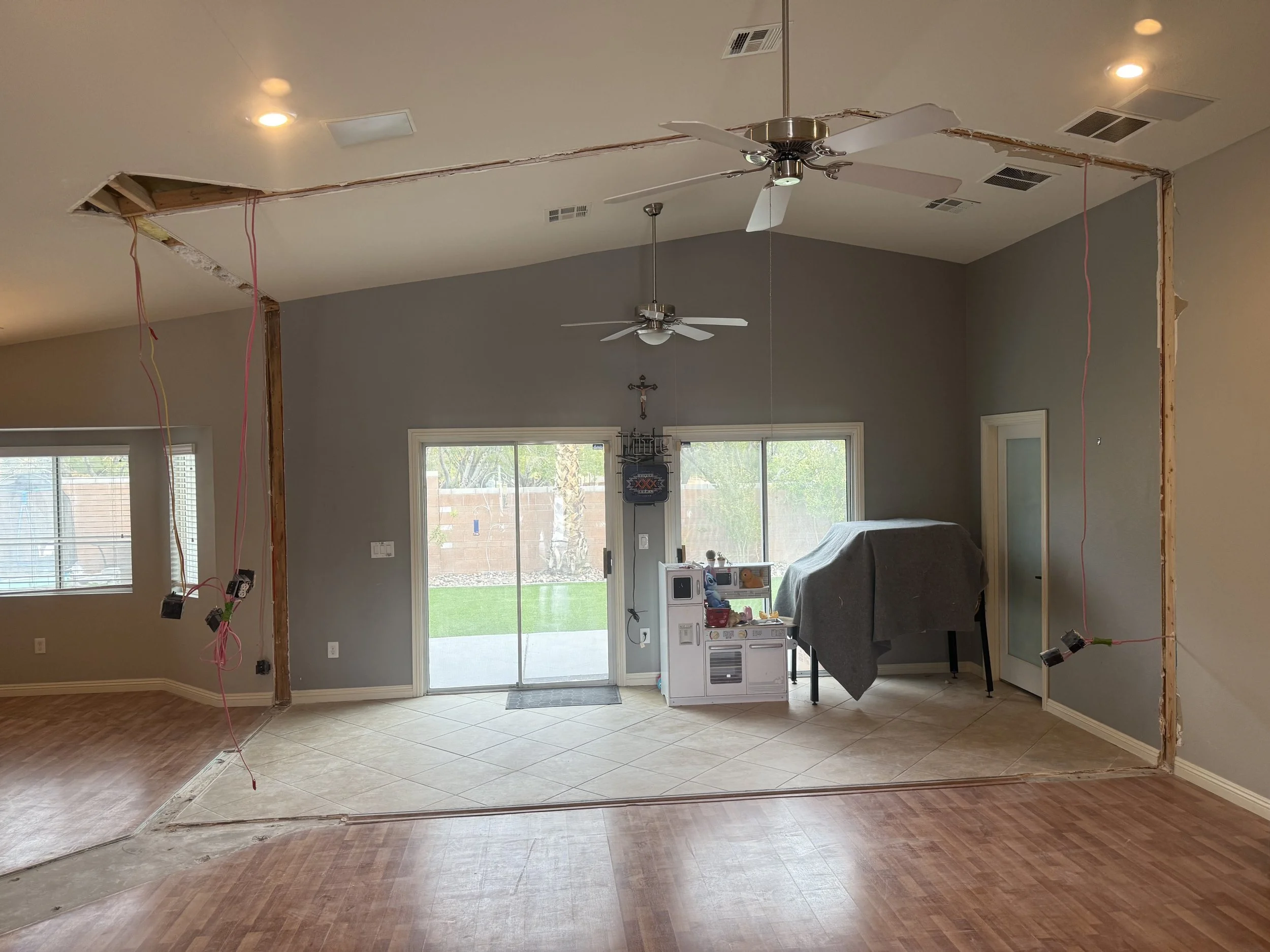 Living room undergoing renovation with an open ceiling showing wiring and framing, ceiling fans, sliding glass doors to backyard, and partial flooring removal.