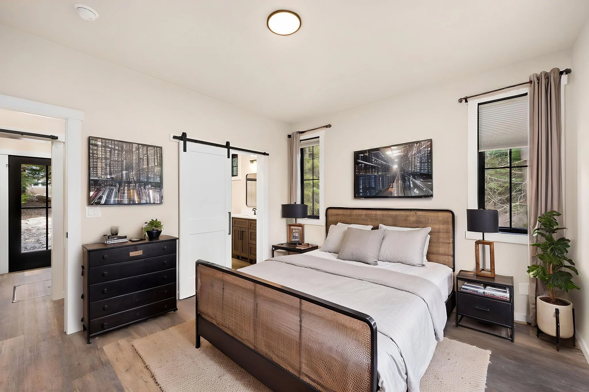 A custom ADU modern bedroom with a large bed, black nightstands, and a flat-screen TV on the wall. There are windows with curtains, and a sliding barn door leads to a bathroom. The room has wood flooring and a simple, cozy decor.