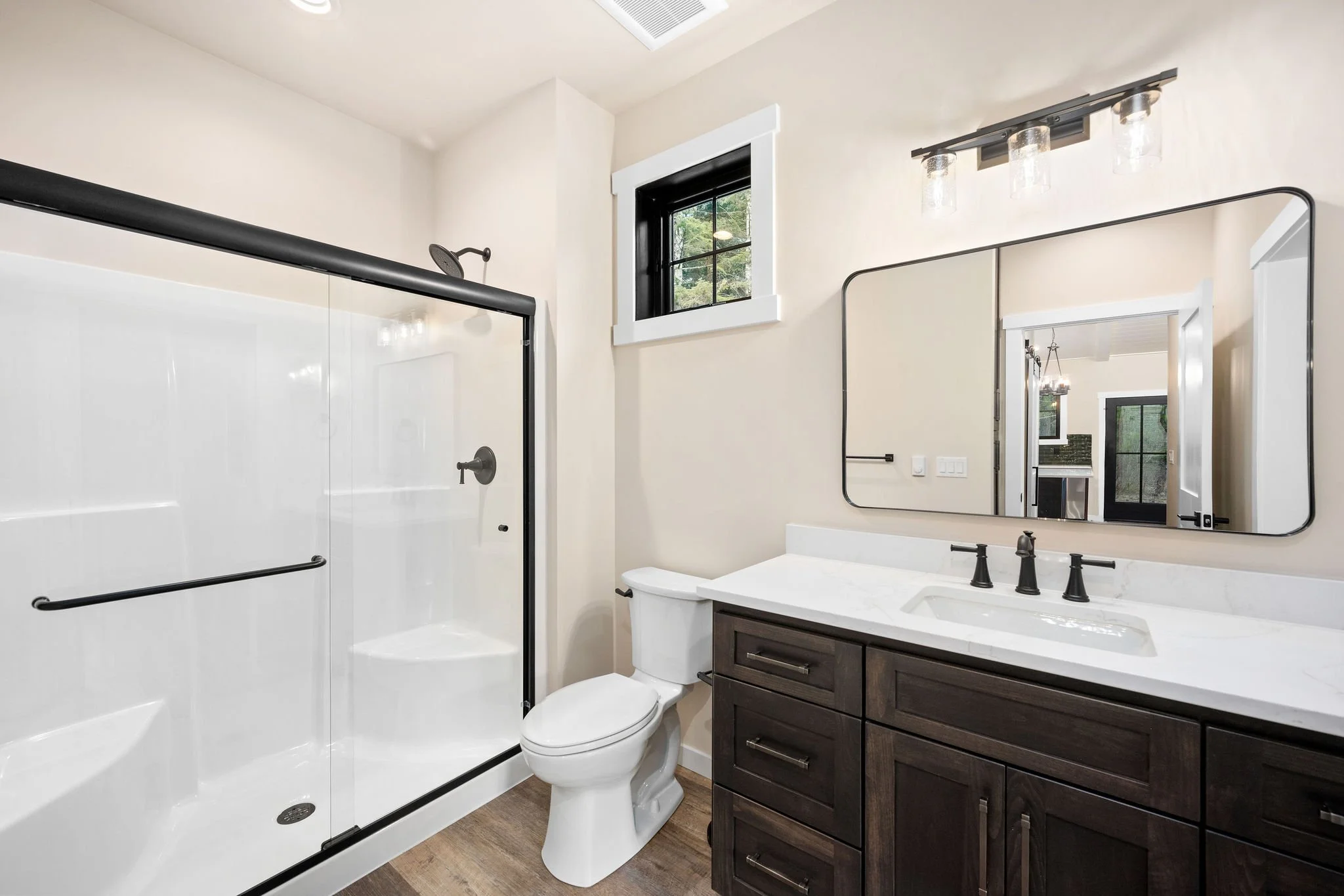 A custom Coeur D'Alene ADU modern bathroom with a walk-in shower with a glass door, a white toilet, and a dark wood vanity with a white countertop and a large mirror. There is a small window with black trim and a black-framed light fixture above the 