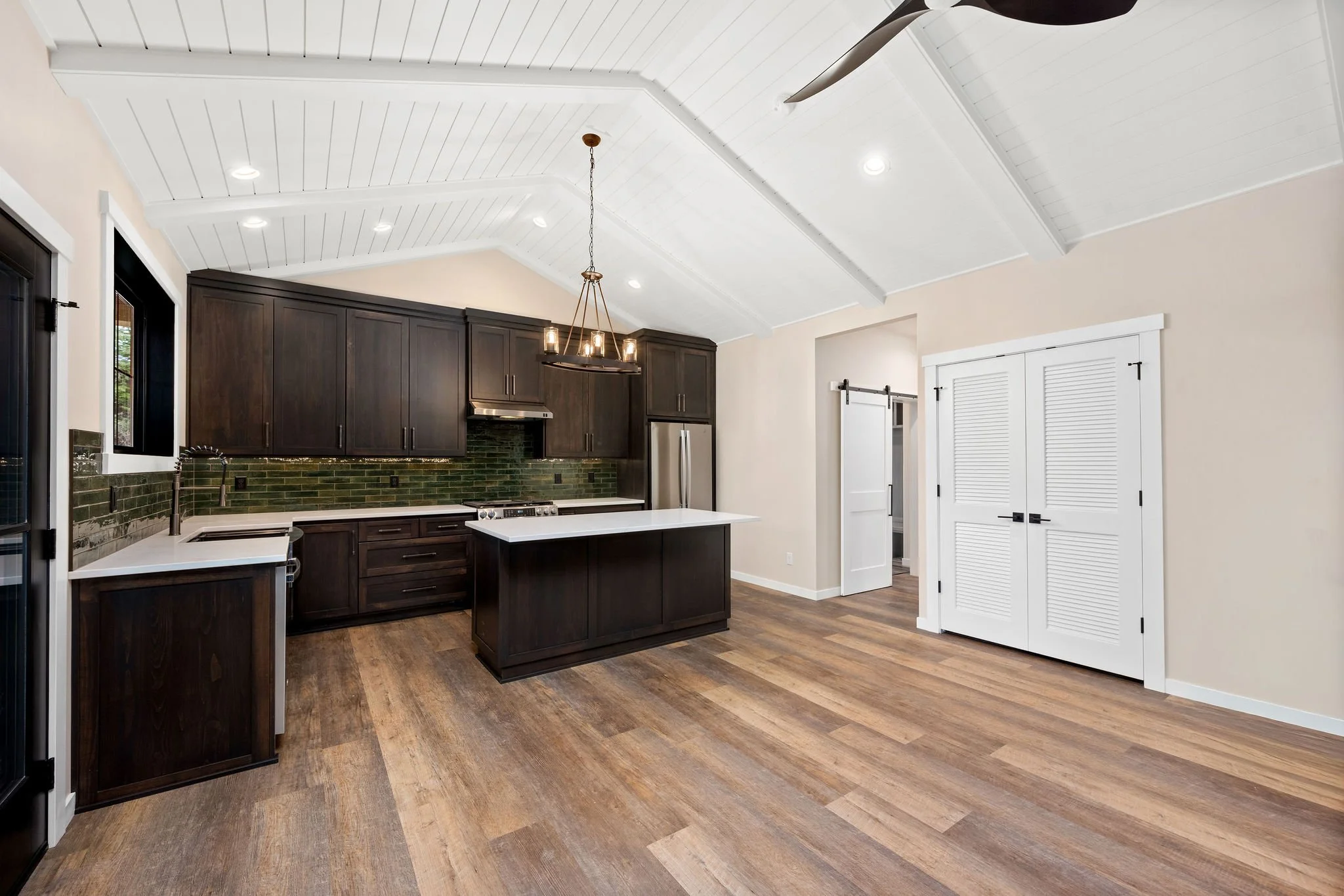 A custom Coeur D'Alene ADU with a modern kitchen with dark wood cabinets, white quartz countertops, green tile backsplash, and a central island. The ceiling is white with recessed lighting and a hanging chandelier. The flooring is wood, and there are