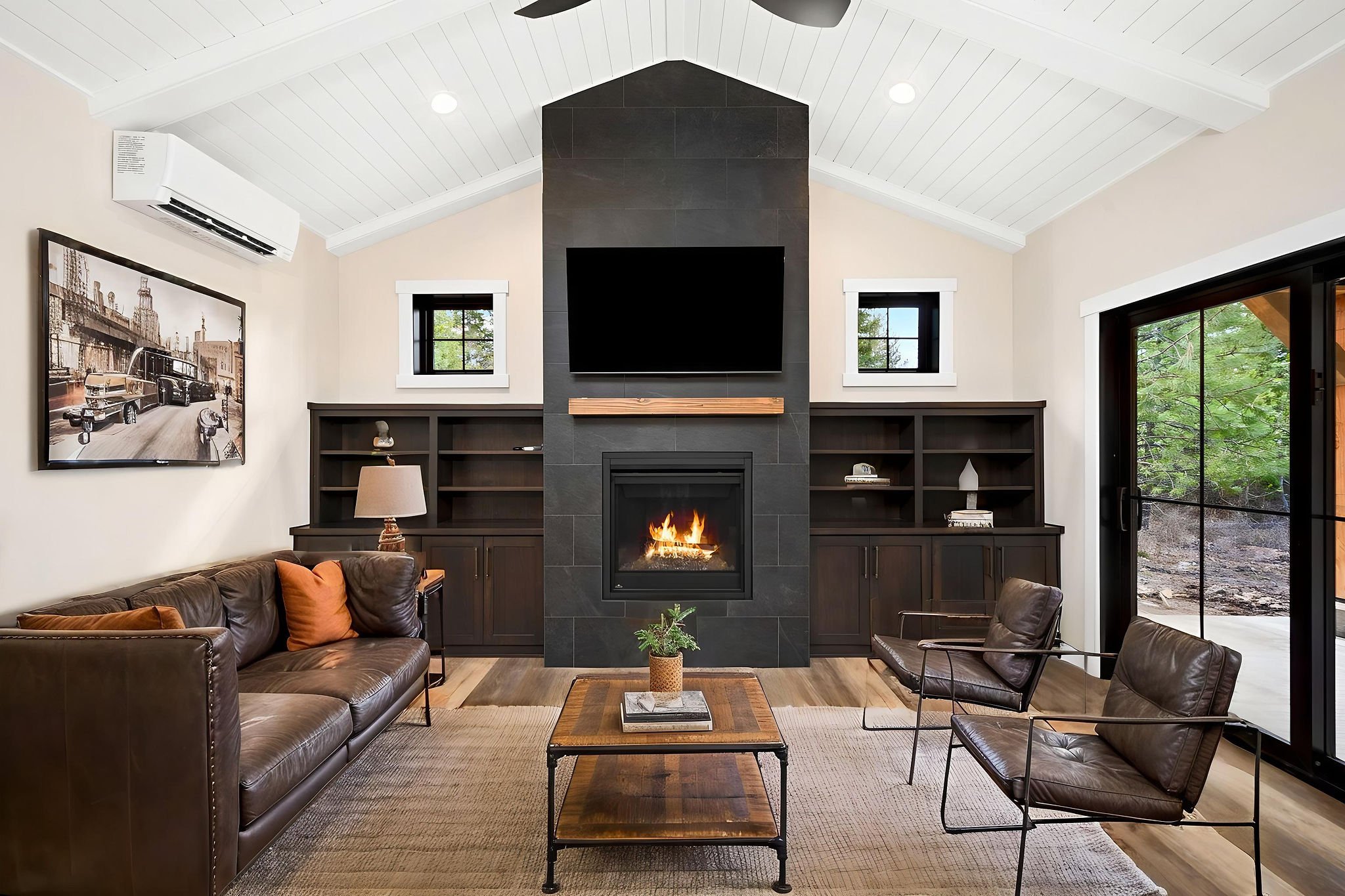 A custom Coeur D'Alene ADU living room with a black stone fireplace, two small windows, built in bookshelves and large sliding glass door, furnished with leather sofas, a wooden coffee table, and a wall-mounted TV.