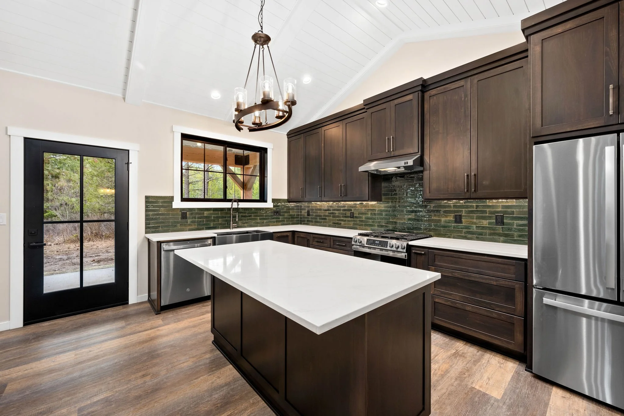Custom ADU Modern kitchen with dark wood cabinets, white countertop island, green tiled backsplash, stainless steel appliances, black door, and large window showing outdoor trees.