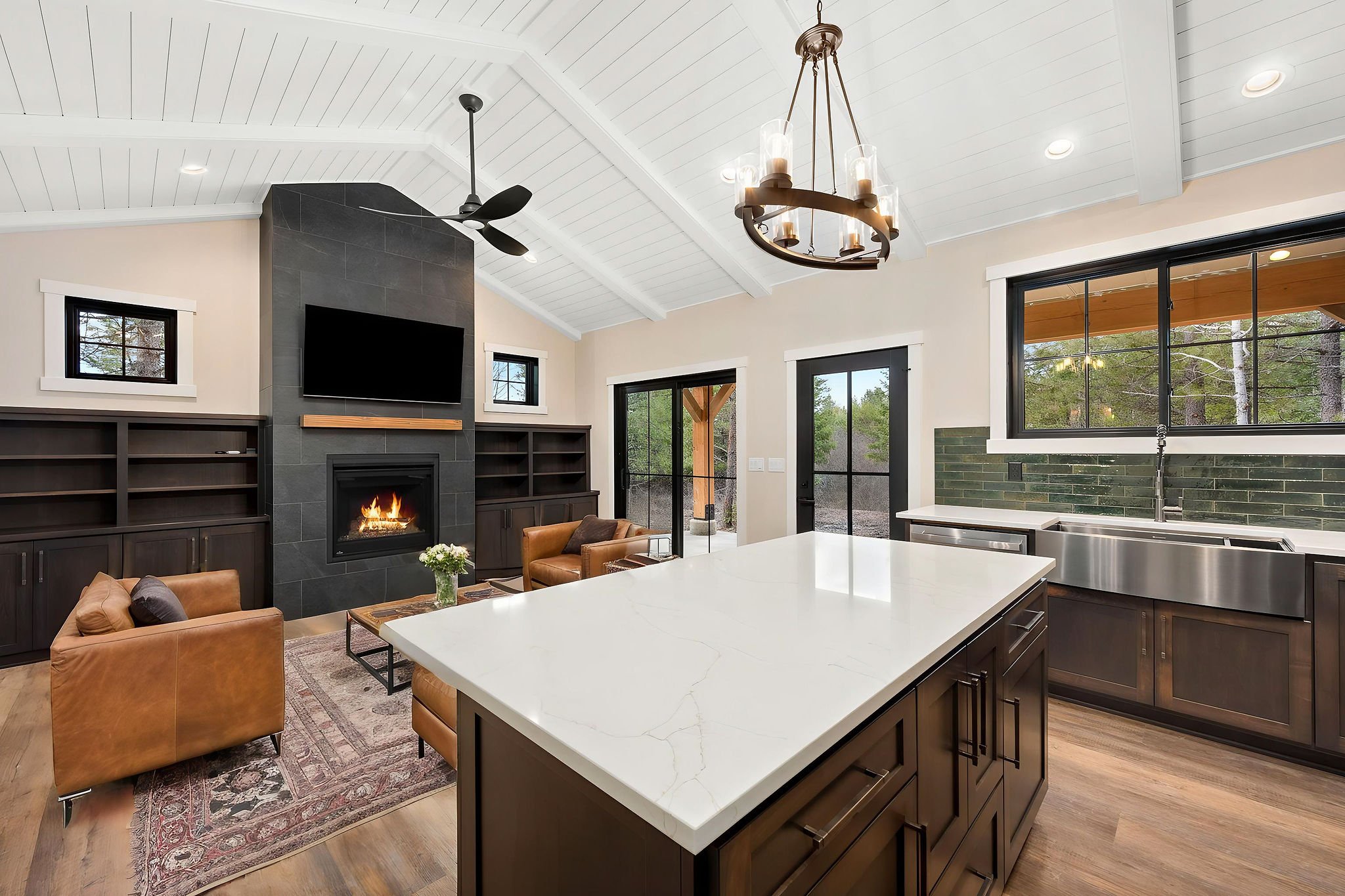 Inside view of a custom Coeur D'Alene ADU with a modern living room with a fireplace, wall-mounted TV, black bookshelves, a large window, and a kitchen island with a white countertop. Includes a ceiling fan and chandelier.
