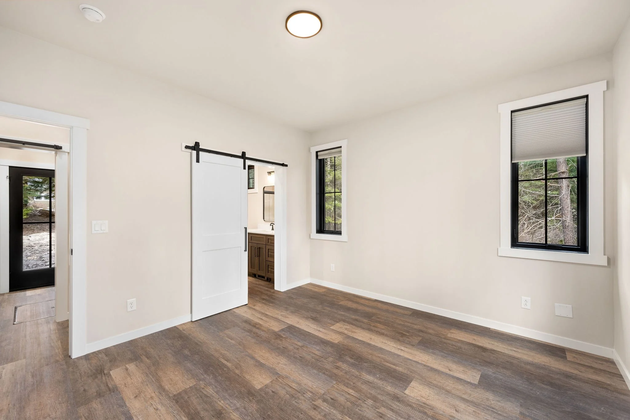 Custom ADU bedroom with white walls, hardwood flooring, two windows with shades, a doorway with a sliding barn door, and a ceiling light.