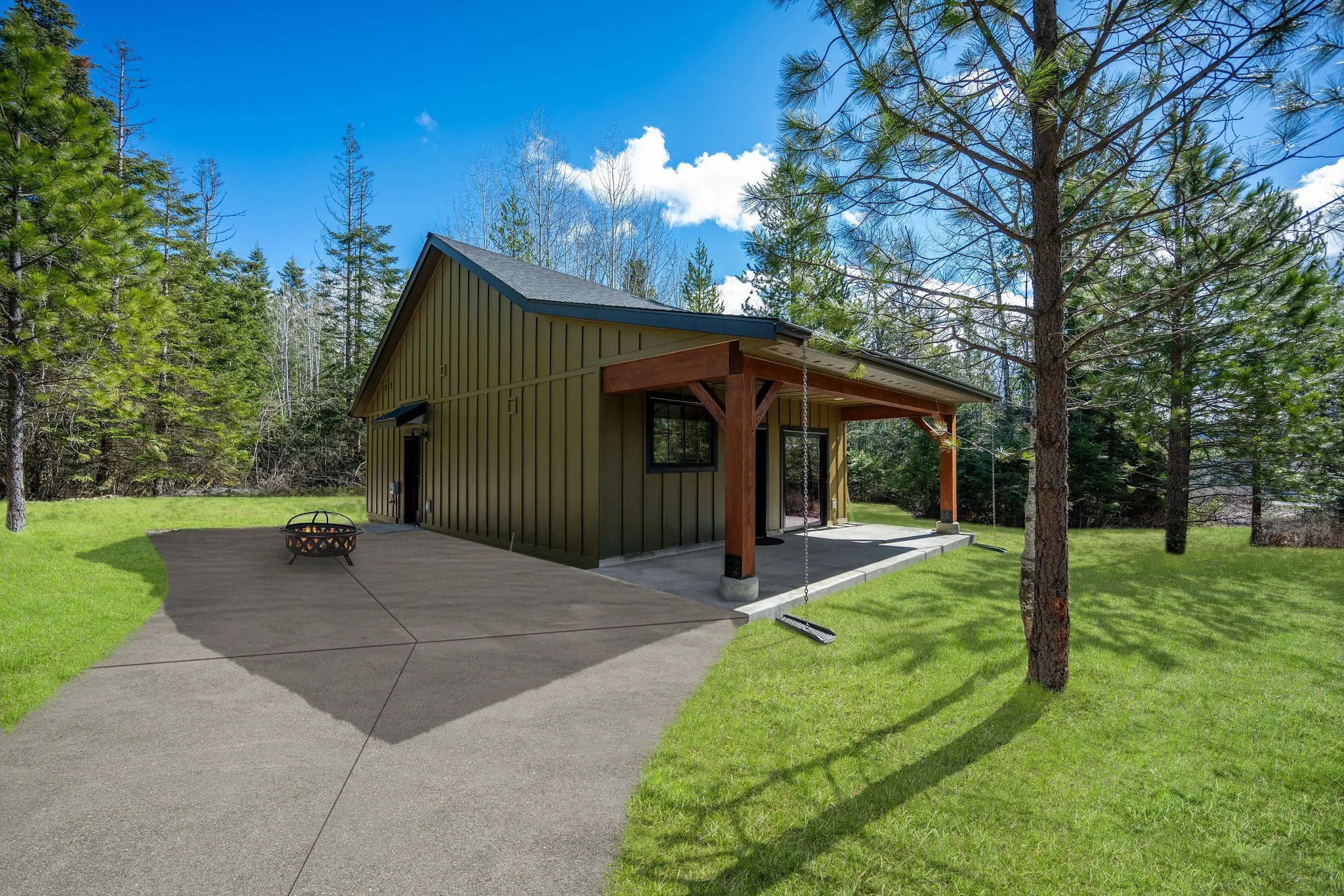 A custom Coeur D'Alene ADU featuring dark green vertical siding and a gray roof, surrounded by tall pine trees under a blue sky with white clouds. There is a concrete patio with a fire pit. 