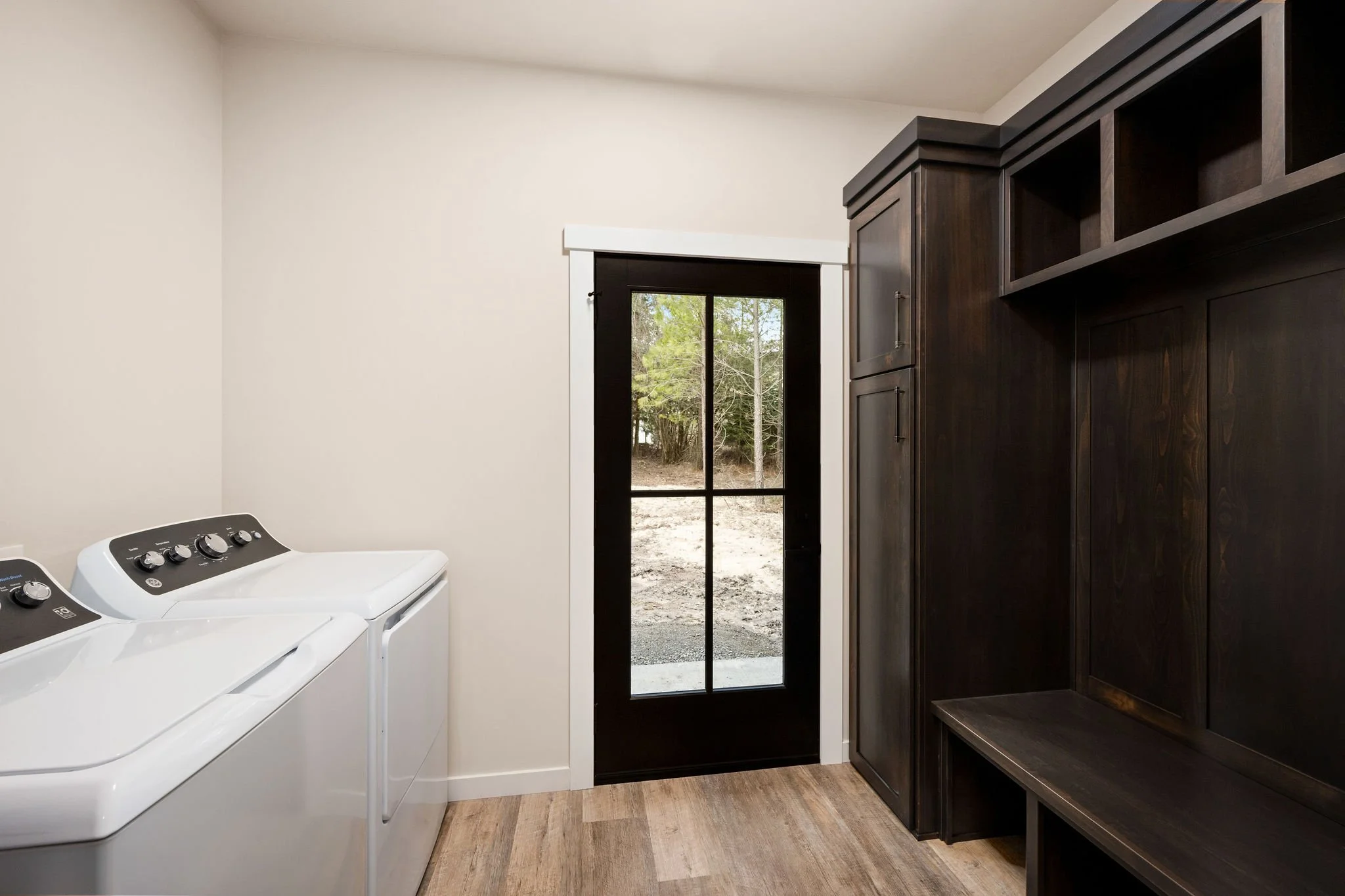 Custom ADU Laundry room with white washing machines, dark wood built in hall tree, and a black door leading outside. 