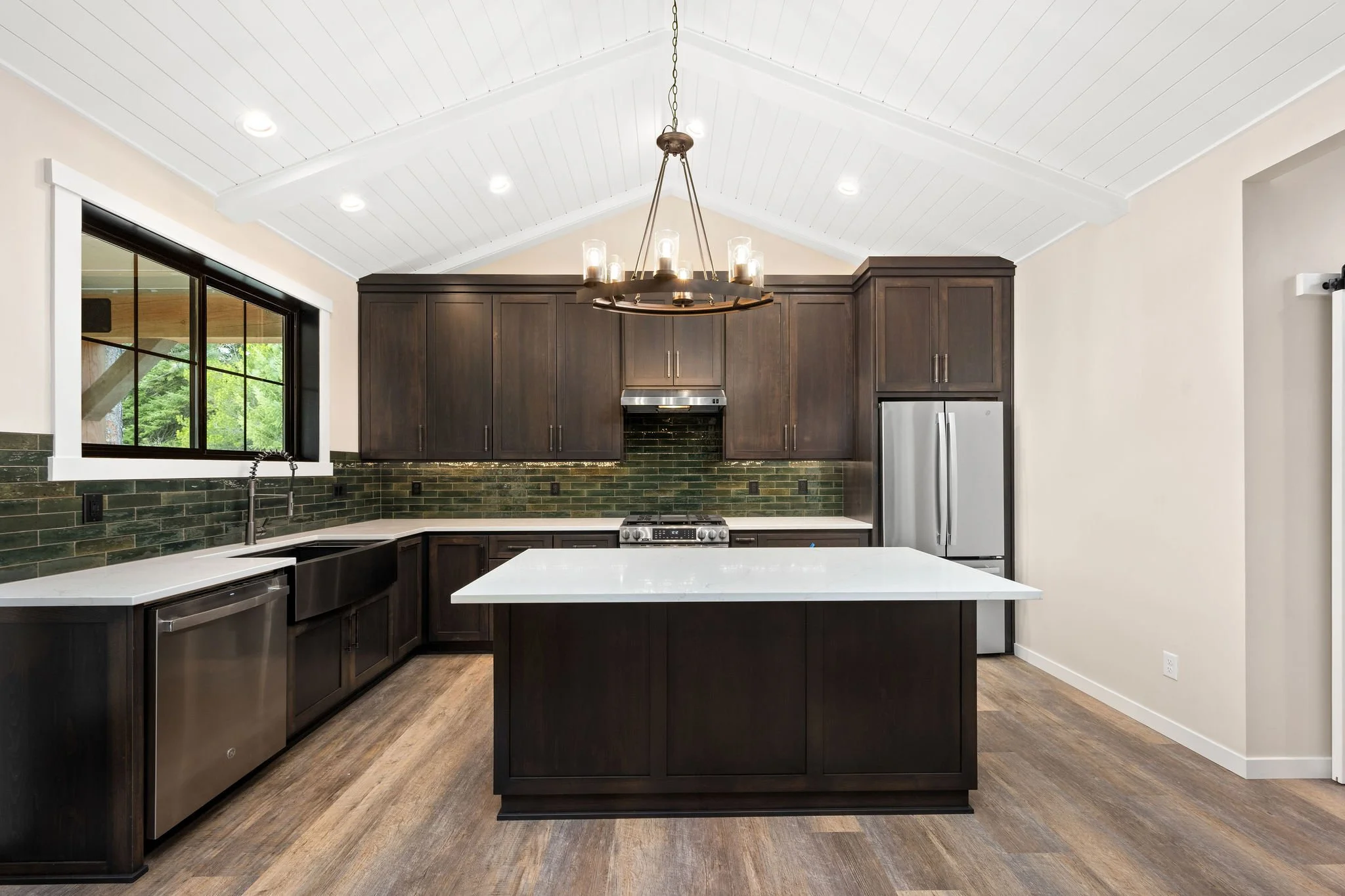 Custom ADU modern kitchen with white counters, dark cabinets, green backsplash tiles, stainless steel appliances, wooden floor, and a black chandelier hanging from a white vaulted ceiling with recessed lighting.