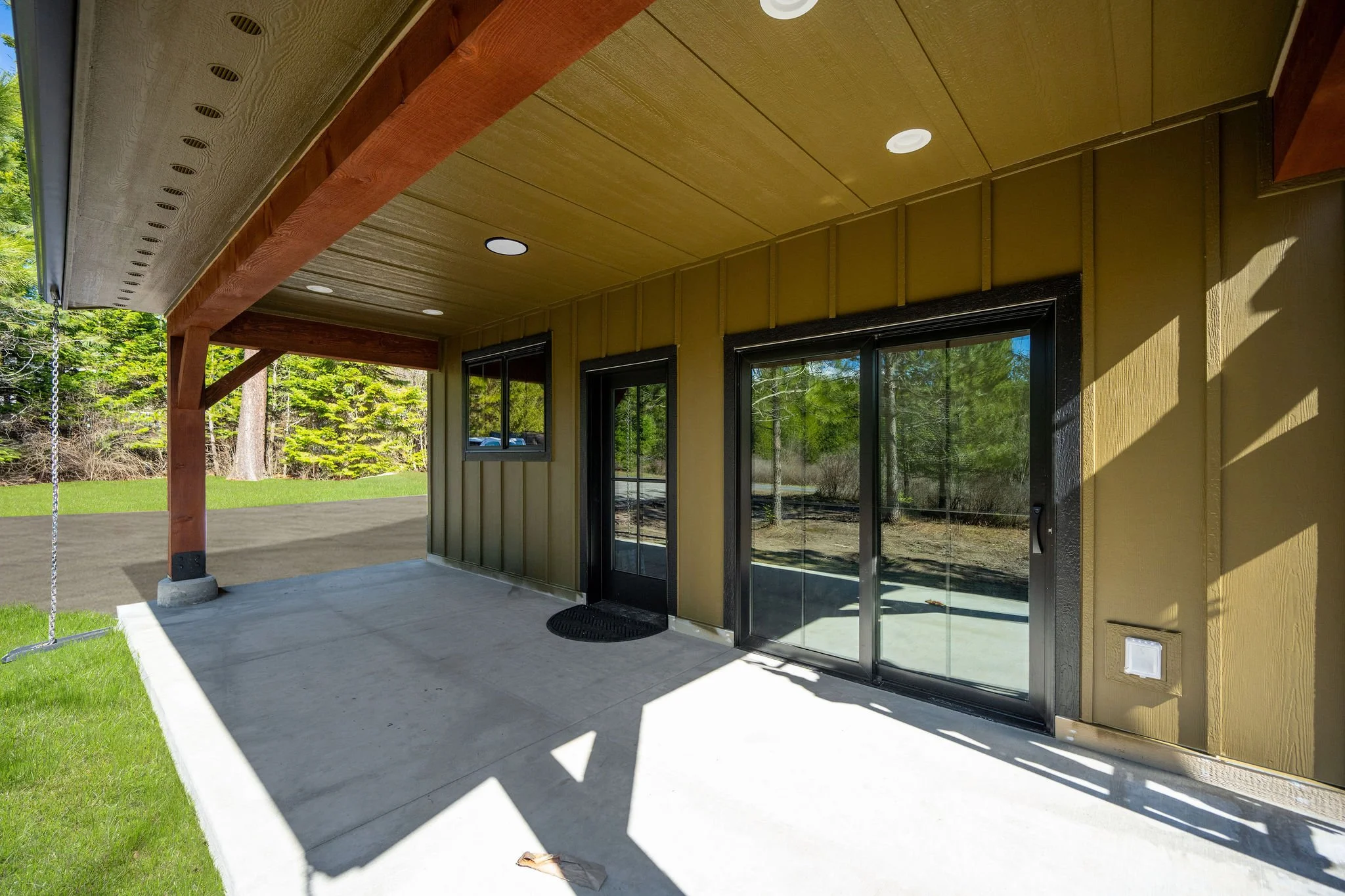 A close up of a Coeur D'Alene ADU covered patio area with a concrete floor, adjacent to a house with green siding and black-framed glass doors. The ceiling is wooden with built-in lights, and there is a swing hanging from the ceiling. 