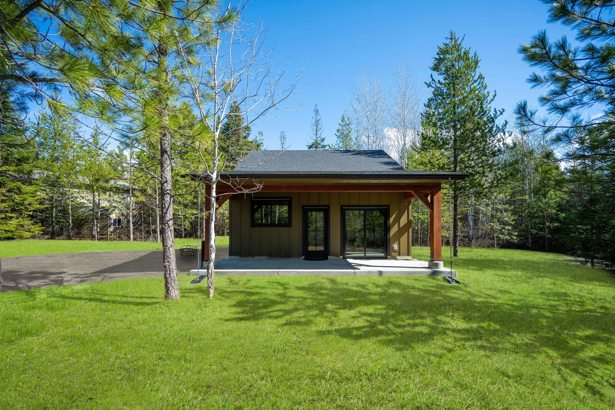 A Coeur D'Alene ADU with large sliding glass doors, situated in a grassy yard surrounded by tall trees, under a blue sky with some clouds.