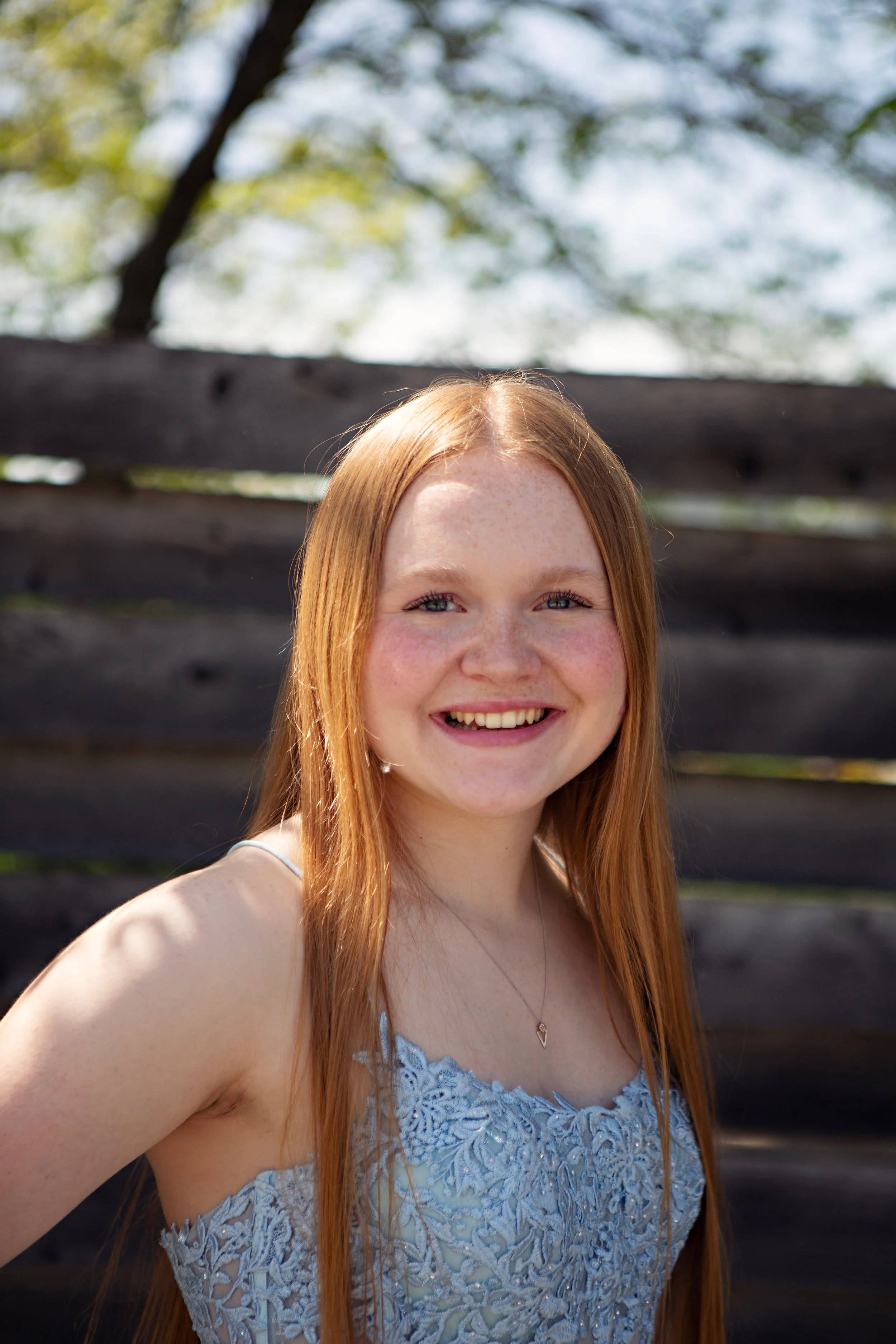 A young woman with long red hair smiling outdoors, wearing a blue lace dress and a heart-shaped necklace, with a wooden fence and trees in the background.