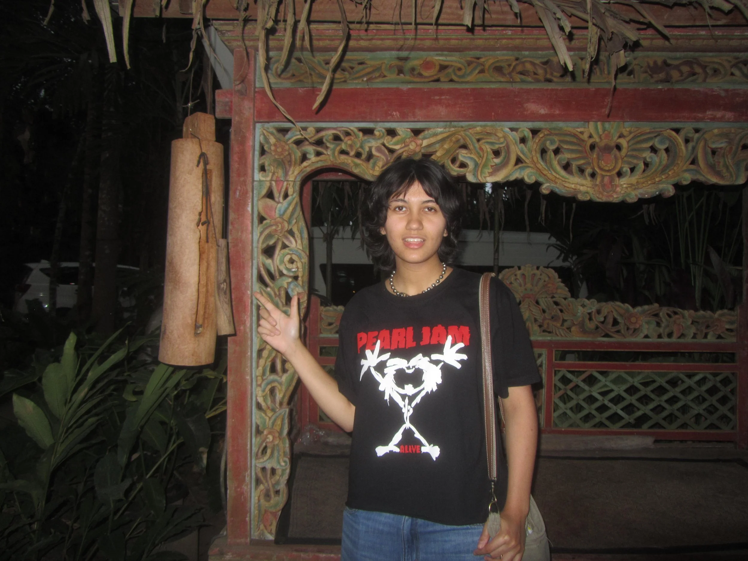 A person with short dark hair wearing a Pearl Jam t-shirt standing in front of a decorative wooden structure at night. The person is pointing towards a sign hanging from the structure that says 'ALIVE' with a design of the map of the world.