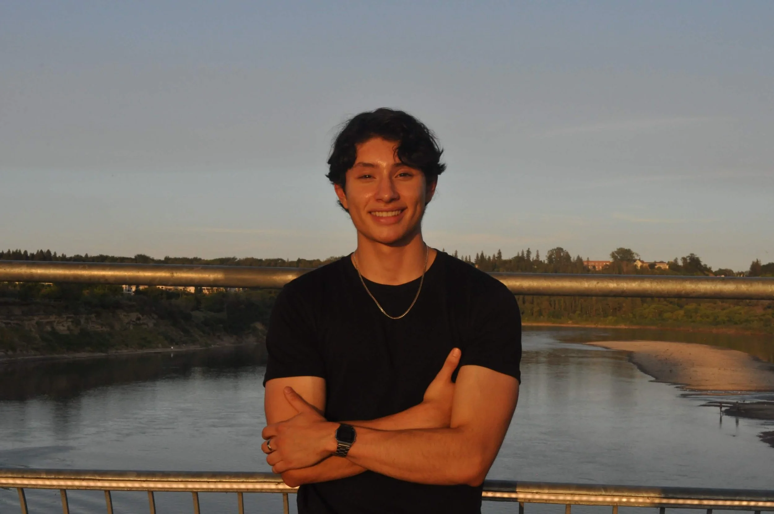 A young man with dark curly hair, smiling, wearing a black shirt, silver chain, watch, and rings, standing with arms crossed in front of a river during sunset, with trees and buildings in the background.