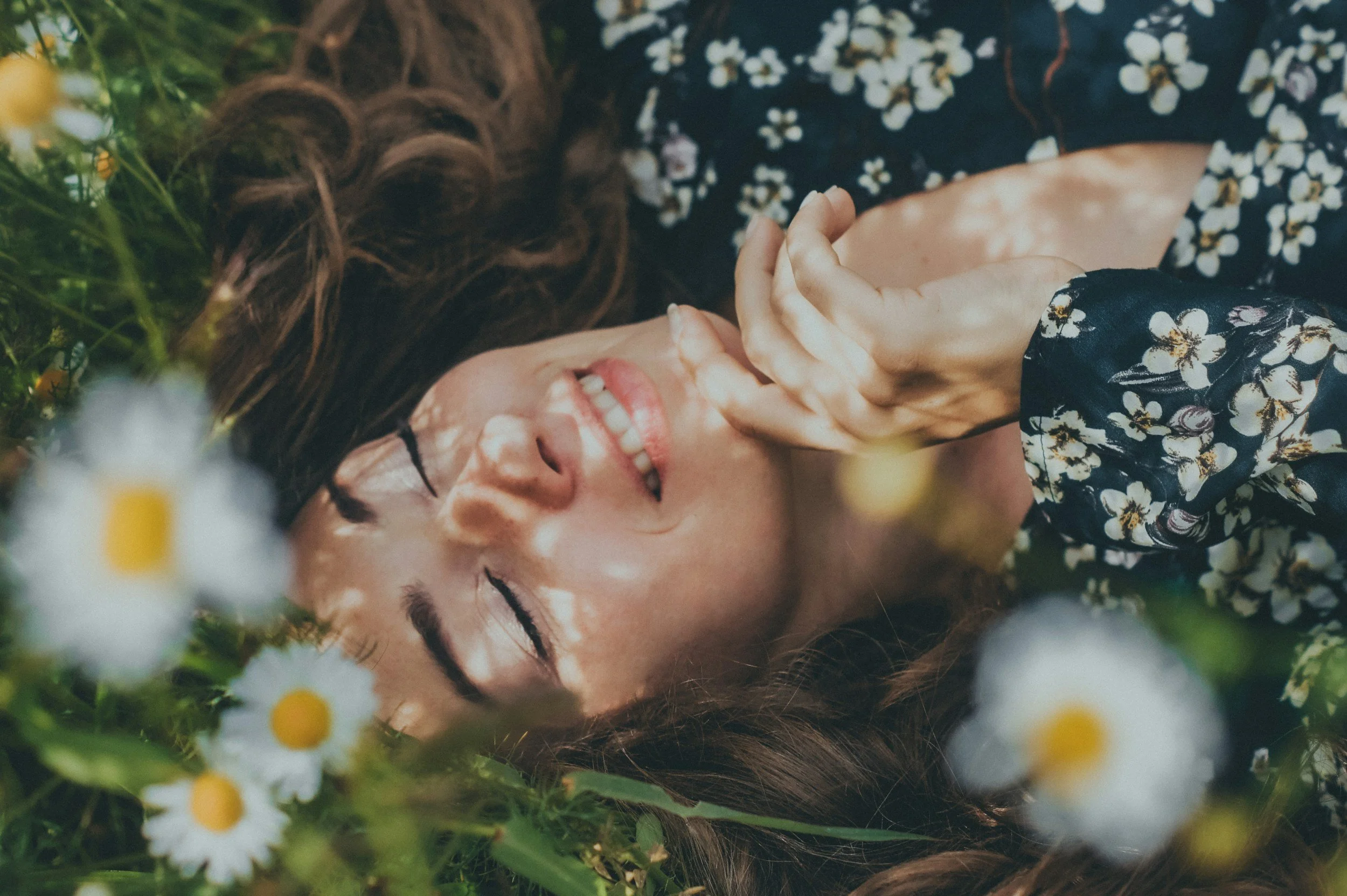 A woman with long brown hair lying on the grass among white daisies with yellow centers, smiling with eyes closed and hand near her face, wearing a dark floral-patterned shirt.