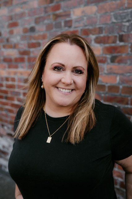A smiling woman with highlighted hair, wearing a black top and gold jewelry, standing against a brick wall.