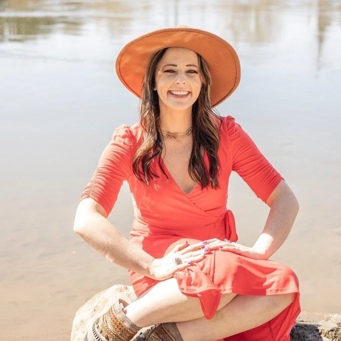 Woman in a red dress and wide-brimmed hat sitting outdoors near water, smiling.