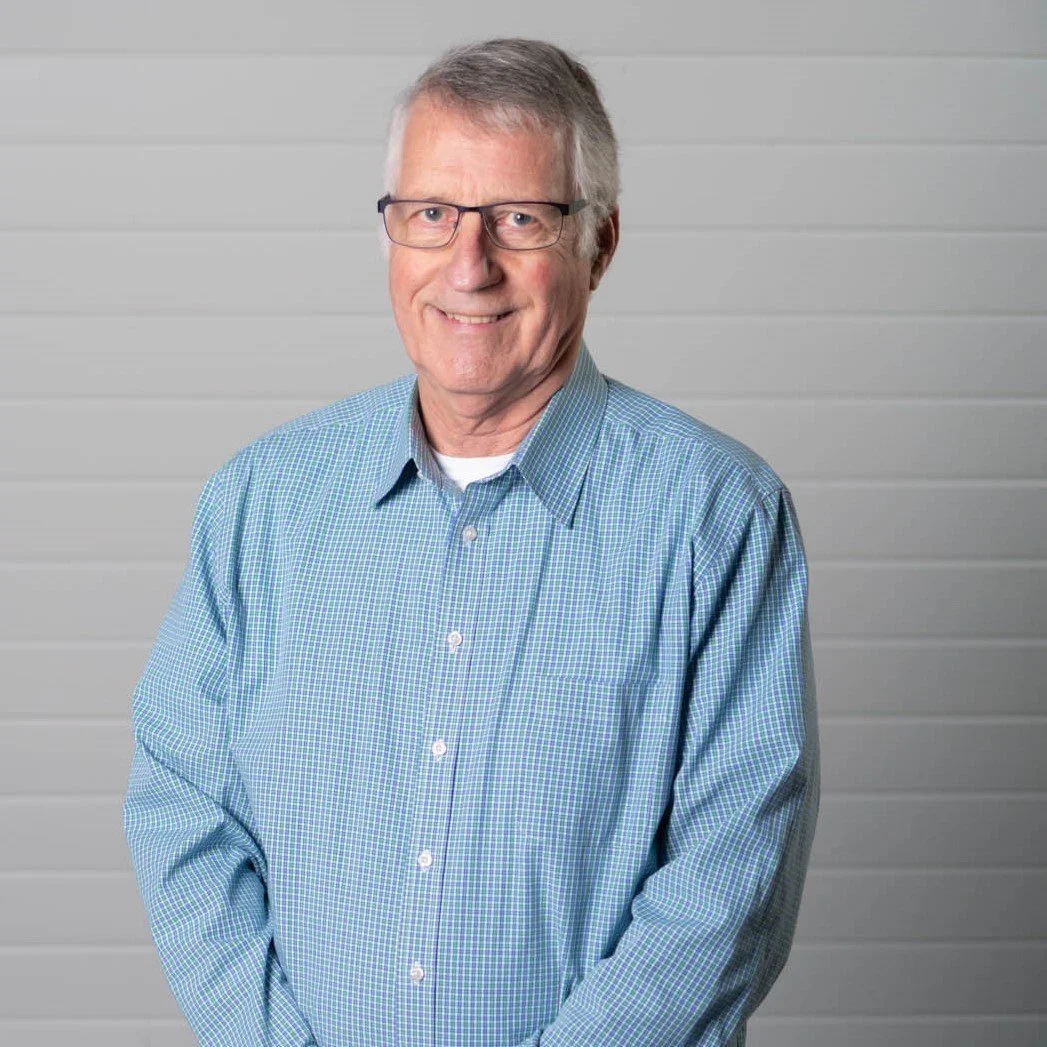 A middle-aged man with glasses, gray hair, and a friendly smile, wearing a blue checkered shirt, standing against a light gray, horizontal paneled background.