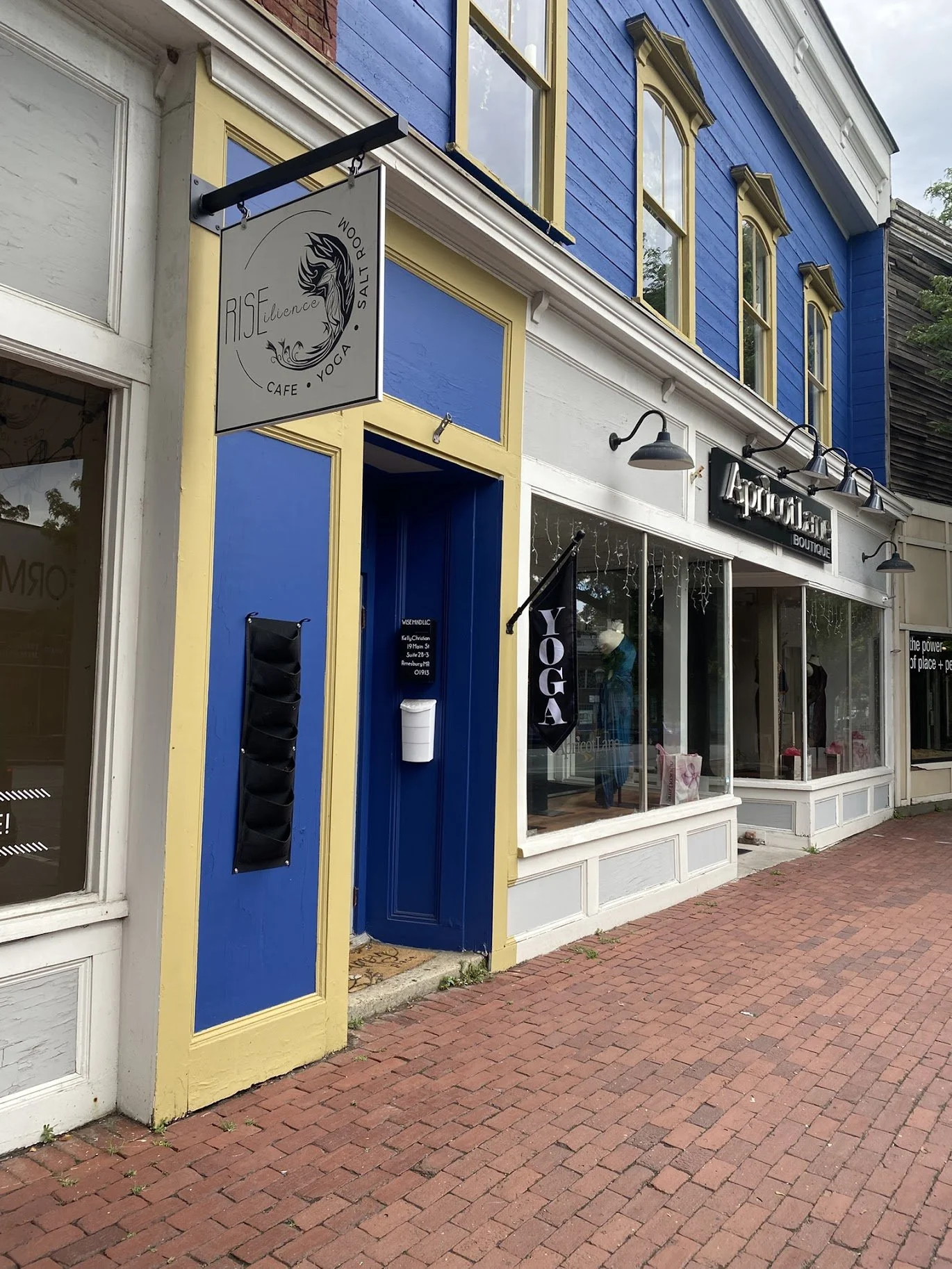Colorful storefront with a sign for Rise Silence Cafe, Yoga, and Salt Room, featuring blue and yellow exterior, large windows with white trim, and a yoga banner outside.