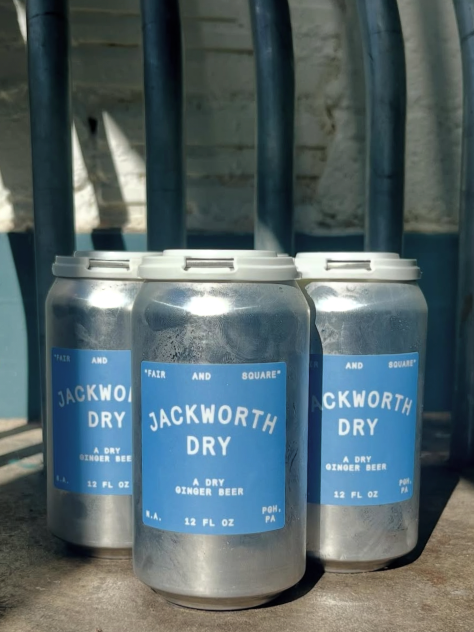 Three cans of Jackworth Dry ginger beer with blue labels on a wooden surface, outdoors with a metal railing in the background.