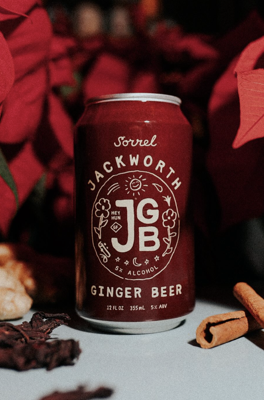 A red can of Jackworth Ginger Beer with alcohol, 12 fluid ounces, 5% ABV, surrounded by red poinsettia leaves, cinnamon sticks, and dried hibiscus flowers.
