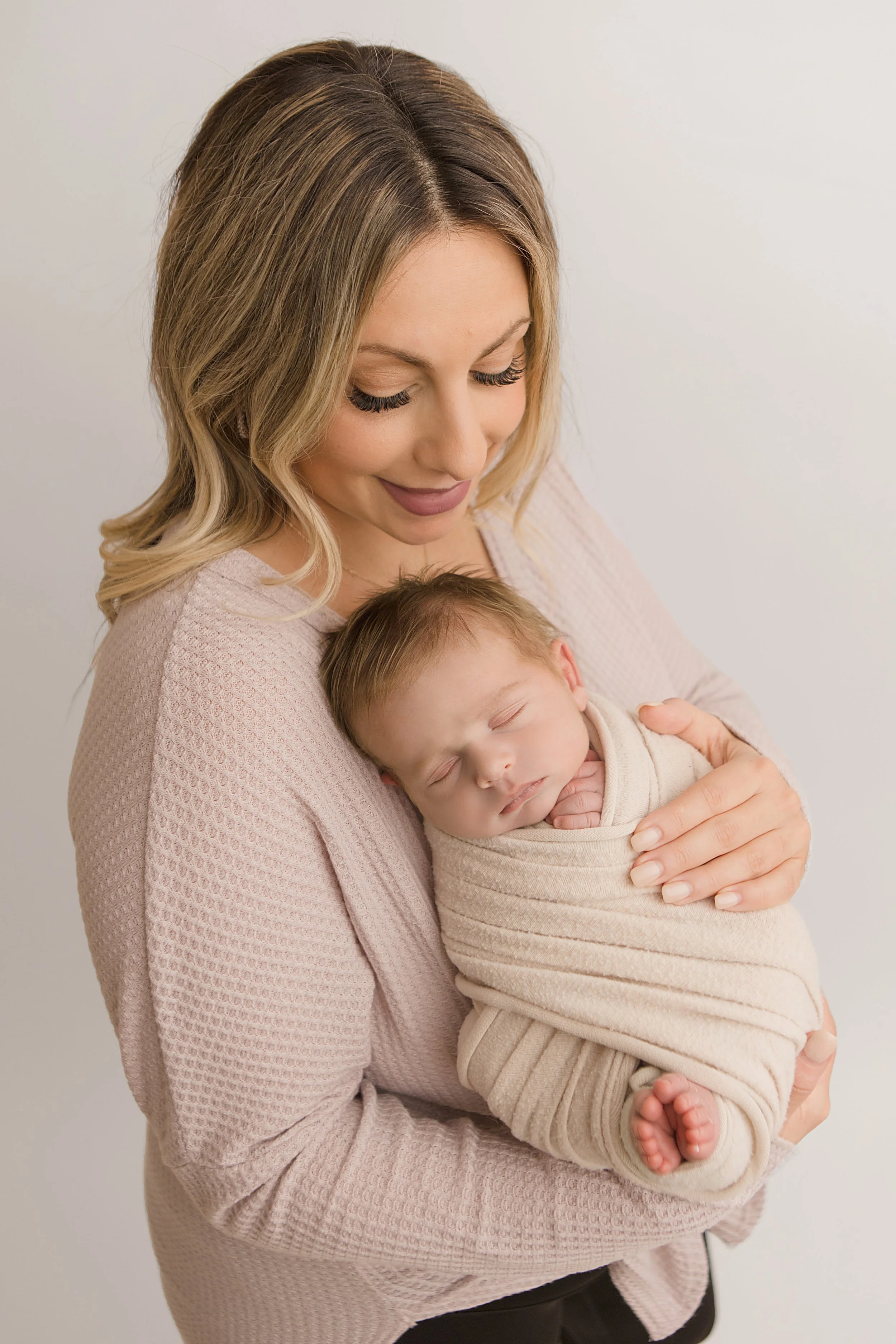 A woman holding a sleeping baby wrapped in beige cloth against a plain white background.