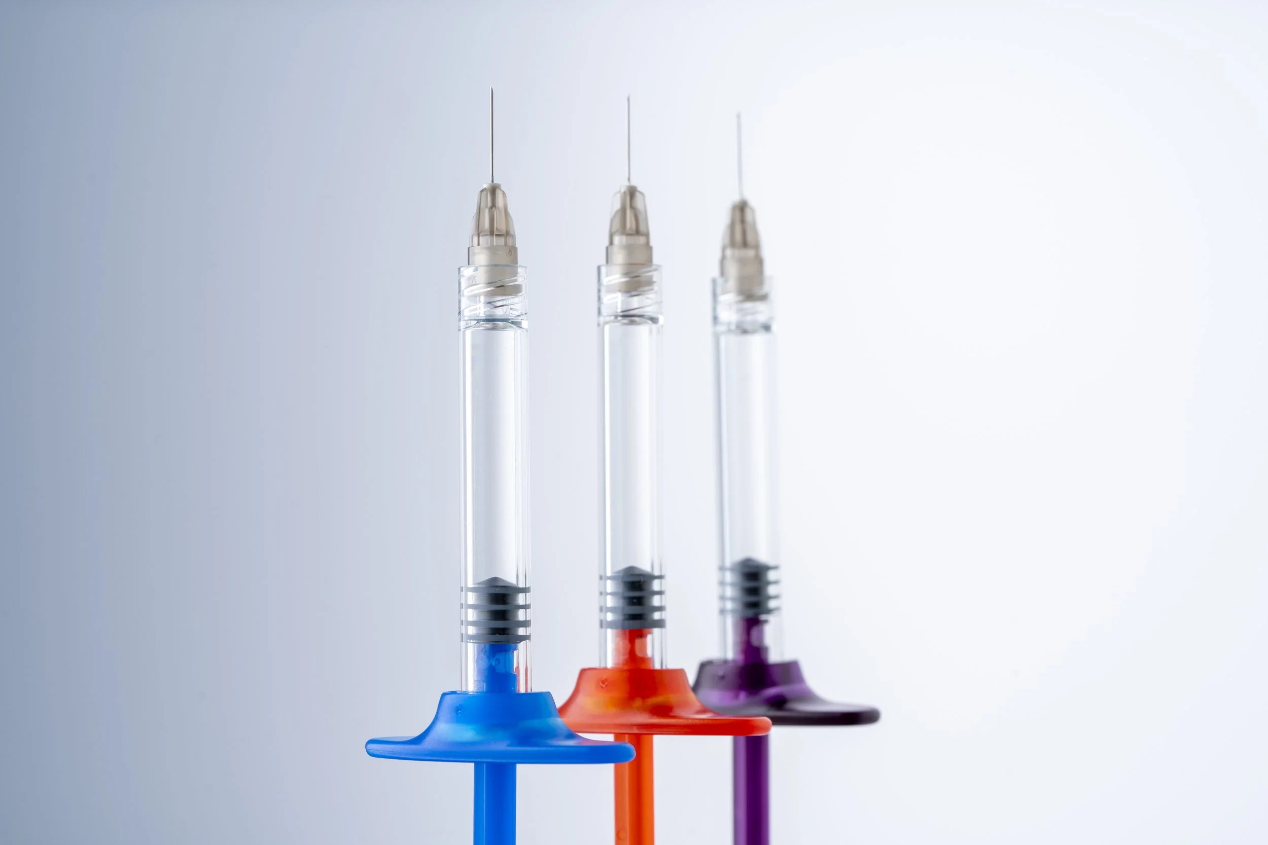 Three syringes filled with clear liquid, each with differently colored bases in blue, orange, and purple, arranged in a row against a plain background.