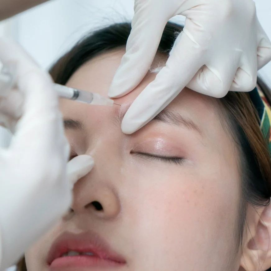 A person receiving a cosmetic injection in the forehead with a syringe, while a medical professional wearing gloves administers the treatment.