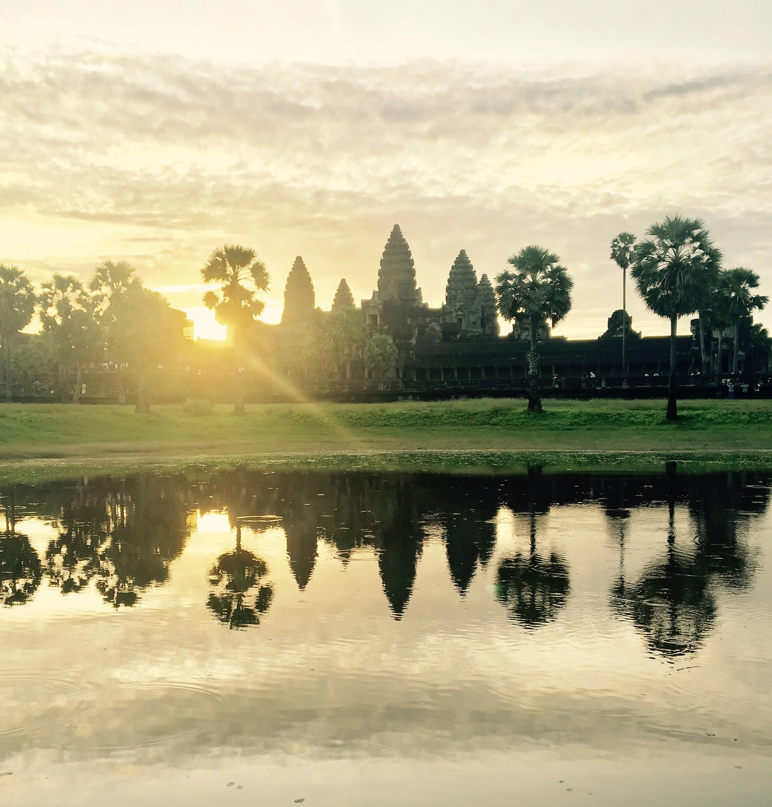 Angkor Wat silhouette at sunrise with reflection in water, surrounded by palm trees.