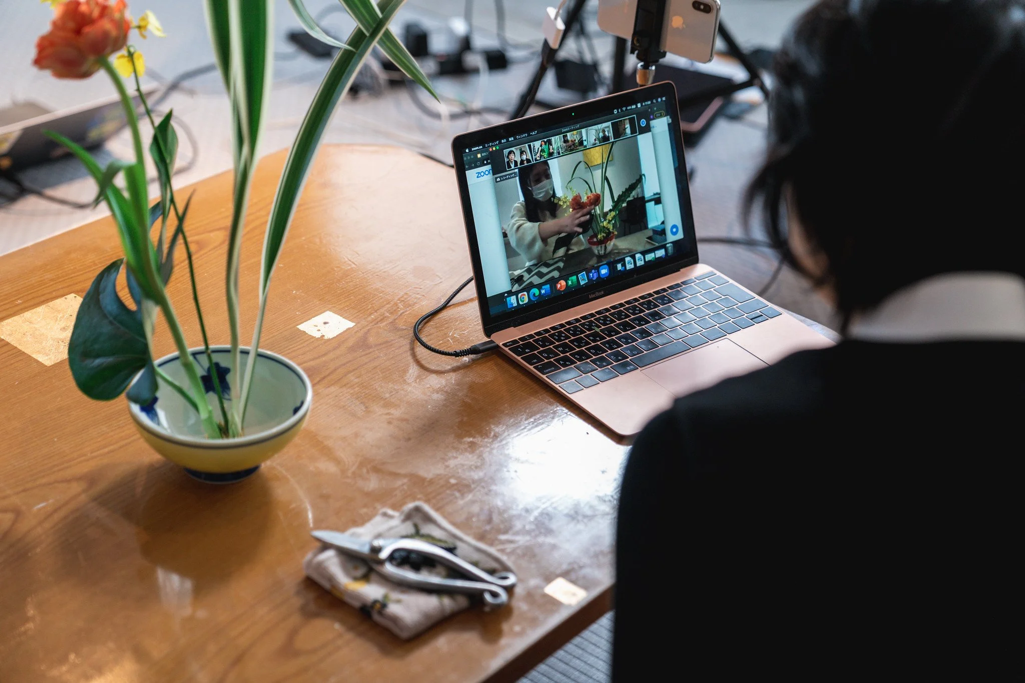 Person participating in an online flower arrangement class via laptop, with a vase of flowers and tools on a wooden table.