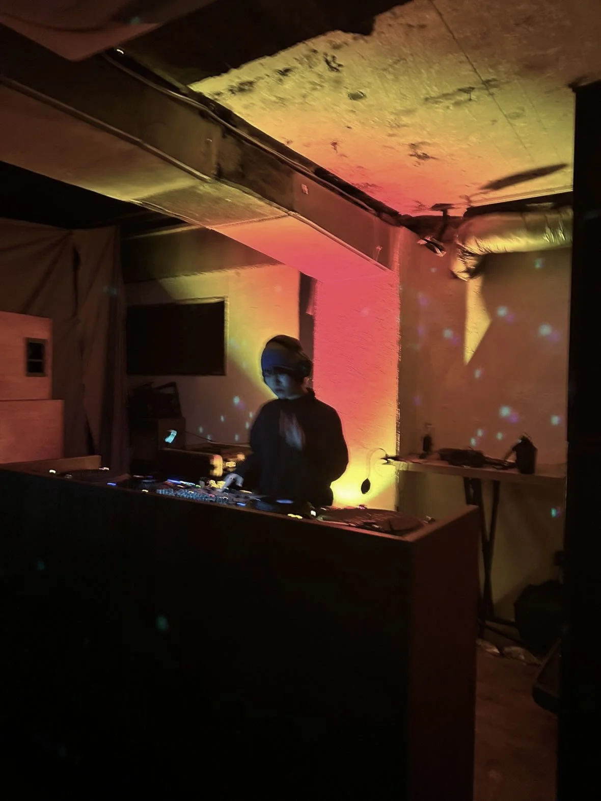 DJ performing in a dimly lit room with colorful lighting, standing behind a DJ booth.
