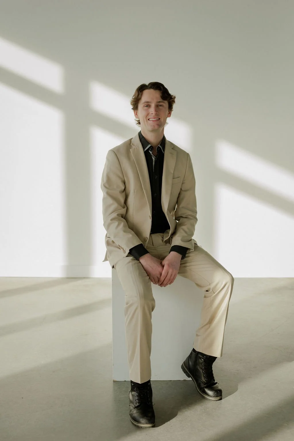 A young man in a beige suit sitting on a white block, smiling, with diagonal sunlight shadows on a plain white background.