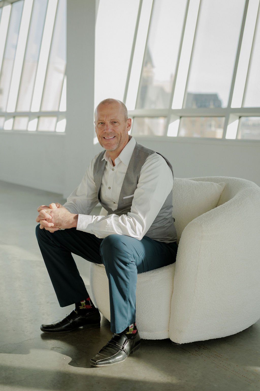 A middle-aged man with a bald head and beard sitting on a white, modern, curved armchair in a bright room with large, angled windowpanes and a cityscape view.
